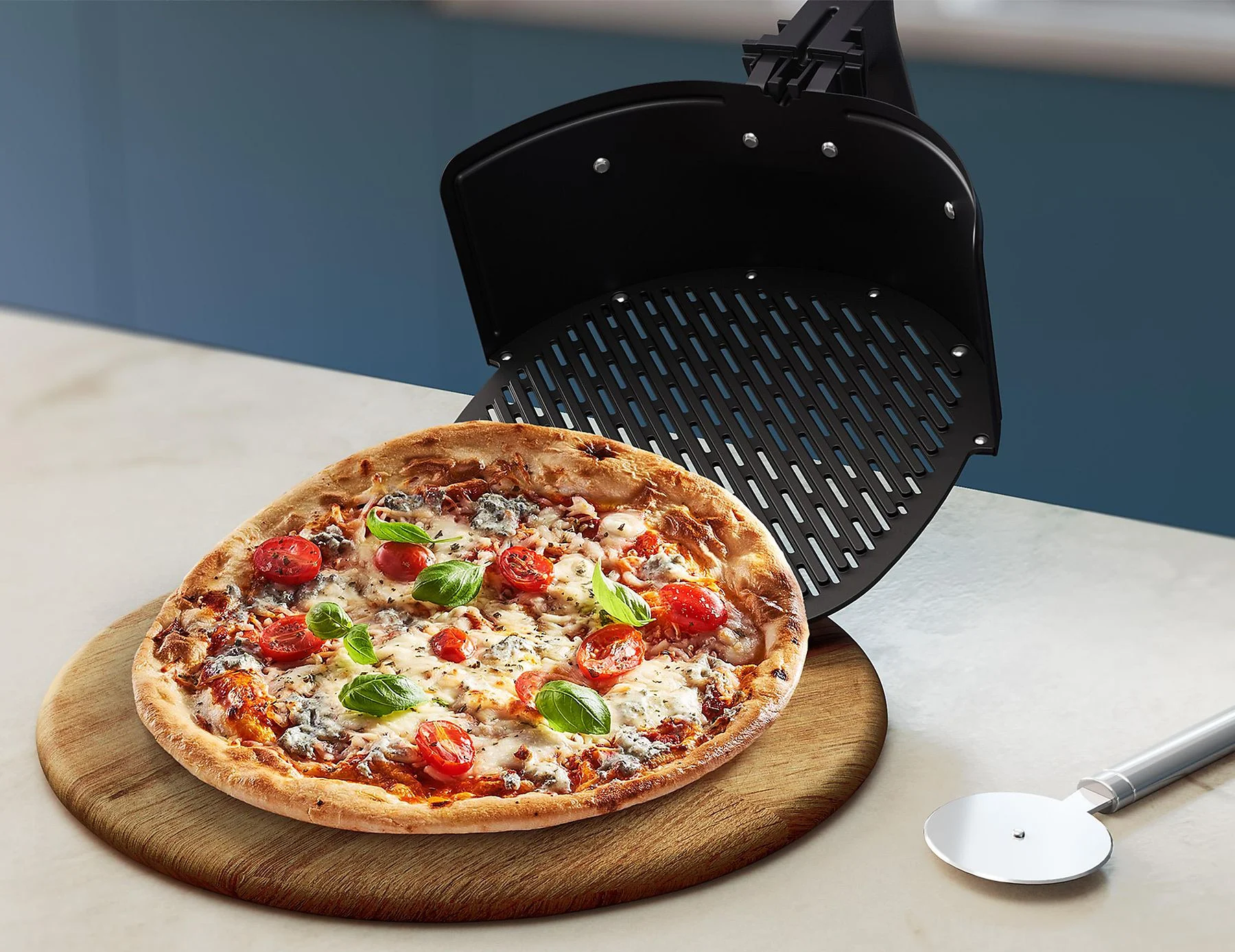 Pizza egy fa deszkán, felette egy air fryer, és egy pizzavágó. Kék fal a háttérben.