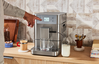 Eine Hand drückt auf eine Taste einer silbernen Kaffeemaschine. Ein Glas befindet sich unter dem Auslauf.