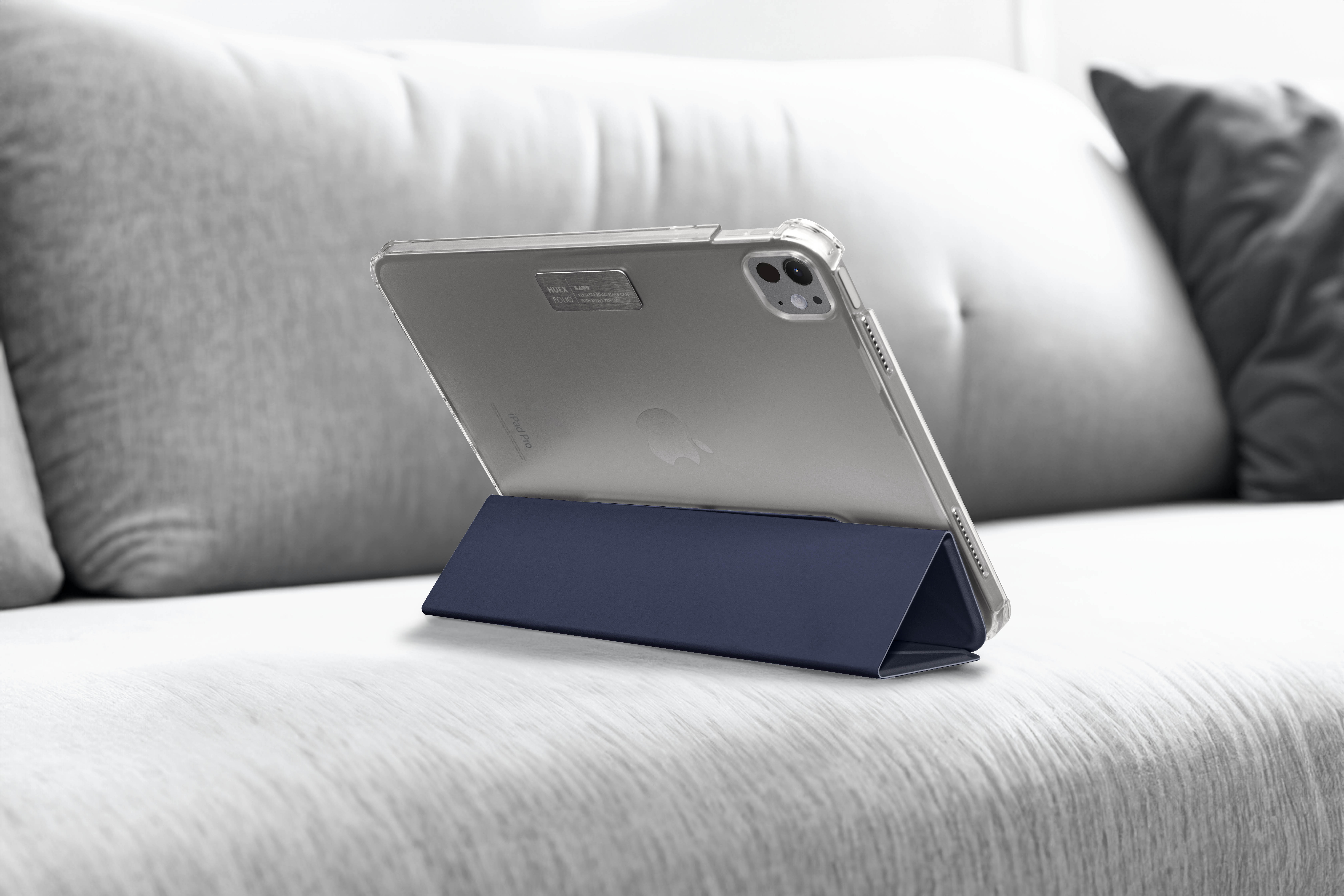 Un iPad in un supporto blu su un divano grigio. L'iPad è in una custodia trasparente. Un cuscino sullo sfondo.