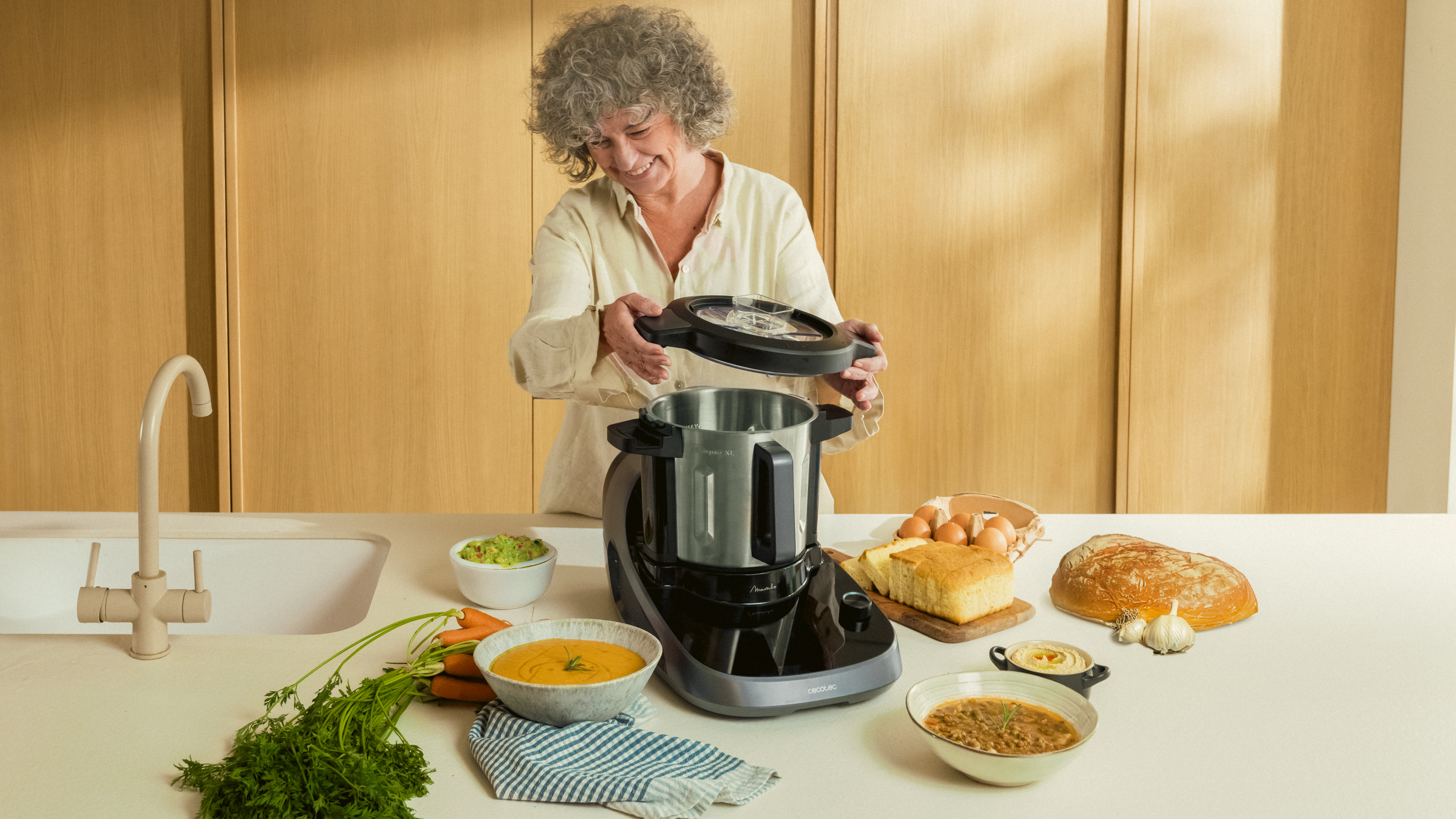 Mujer preparando comida con un electrodoméstico de cocina, rodeada de ingredientes en una encimera.