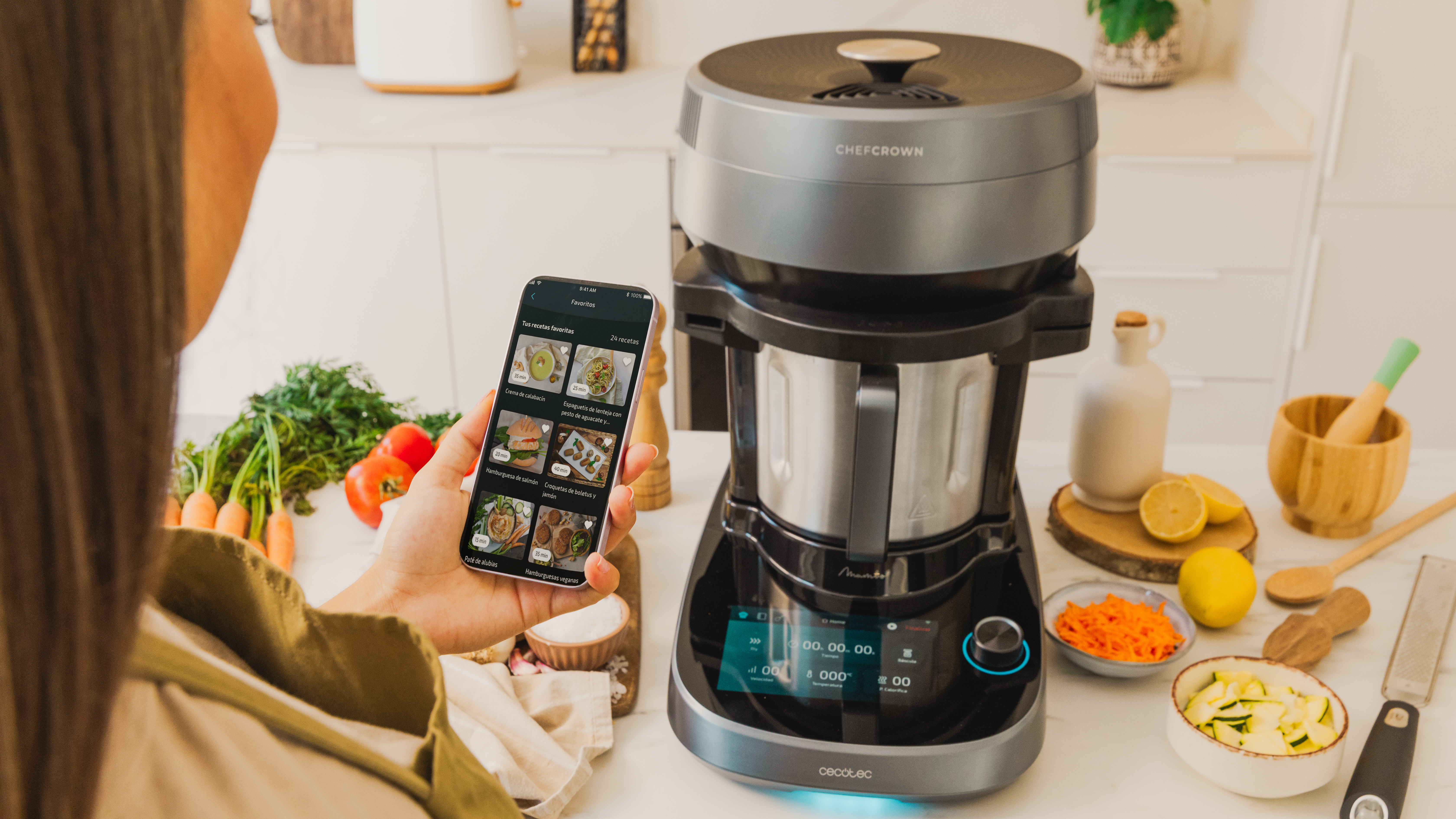 Mujer sosteniendo un teléfono que muestra recetas, con un procesador de alimentos e ingredientes en la encimera.