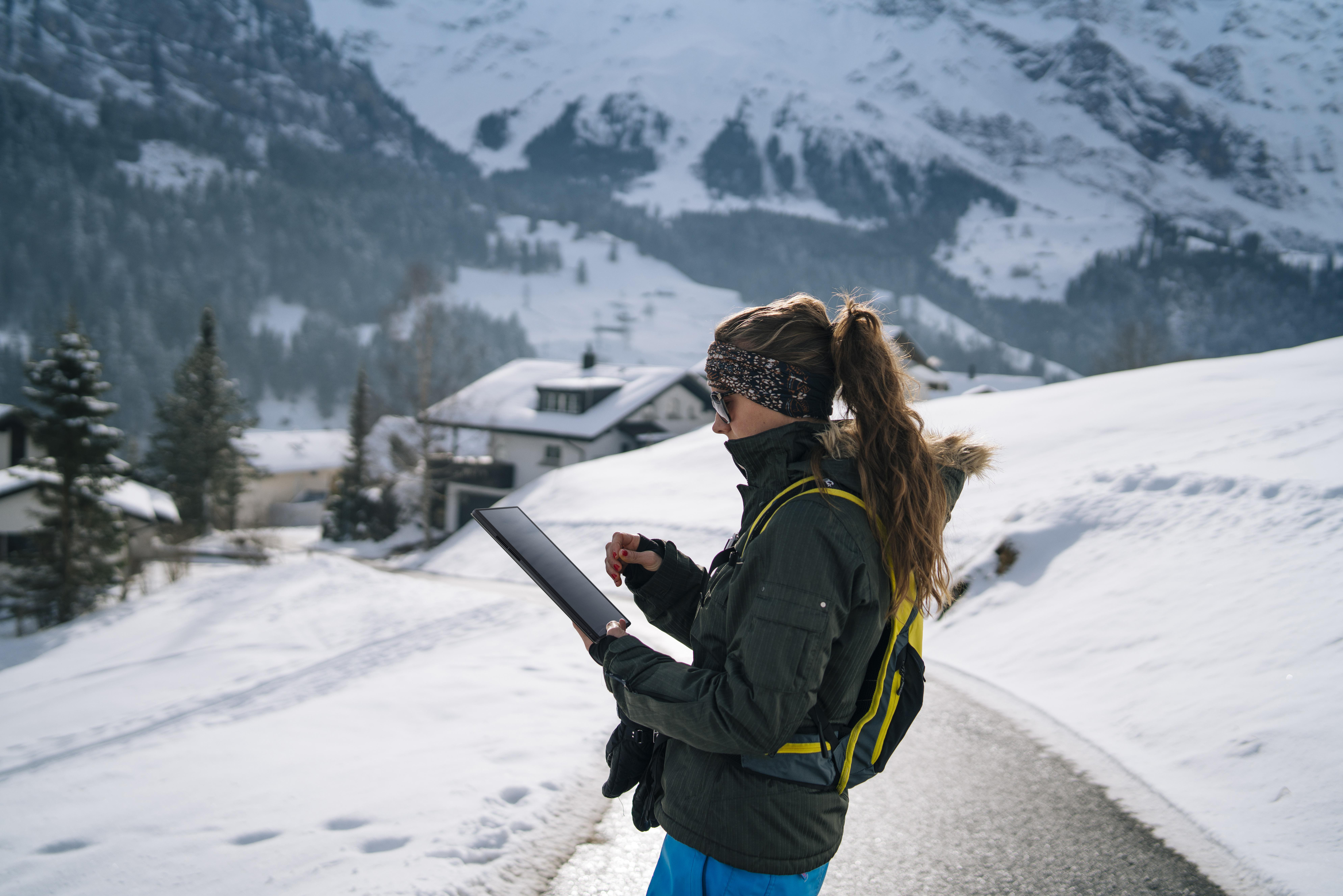 Frau in Winterkleidung, hält Tablet, steht auf schneebedeckter Straße. Berge und Gebäude im Hintergrund.