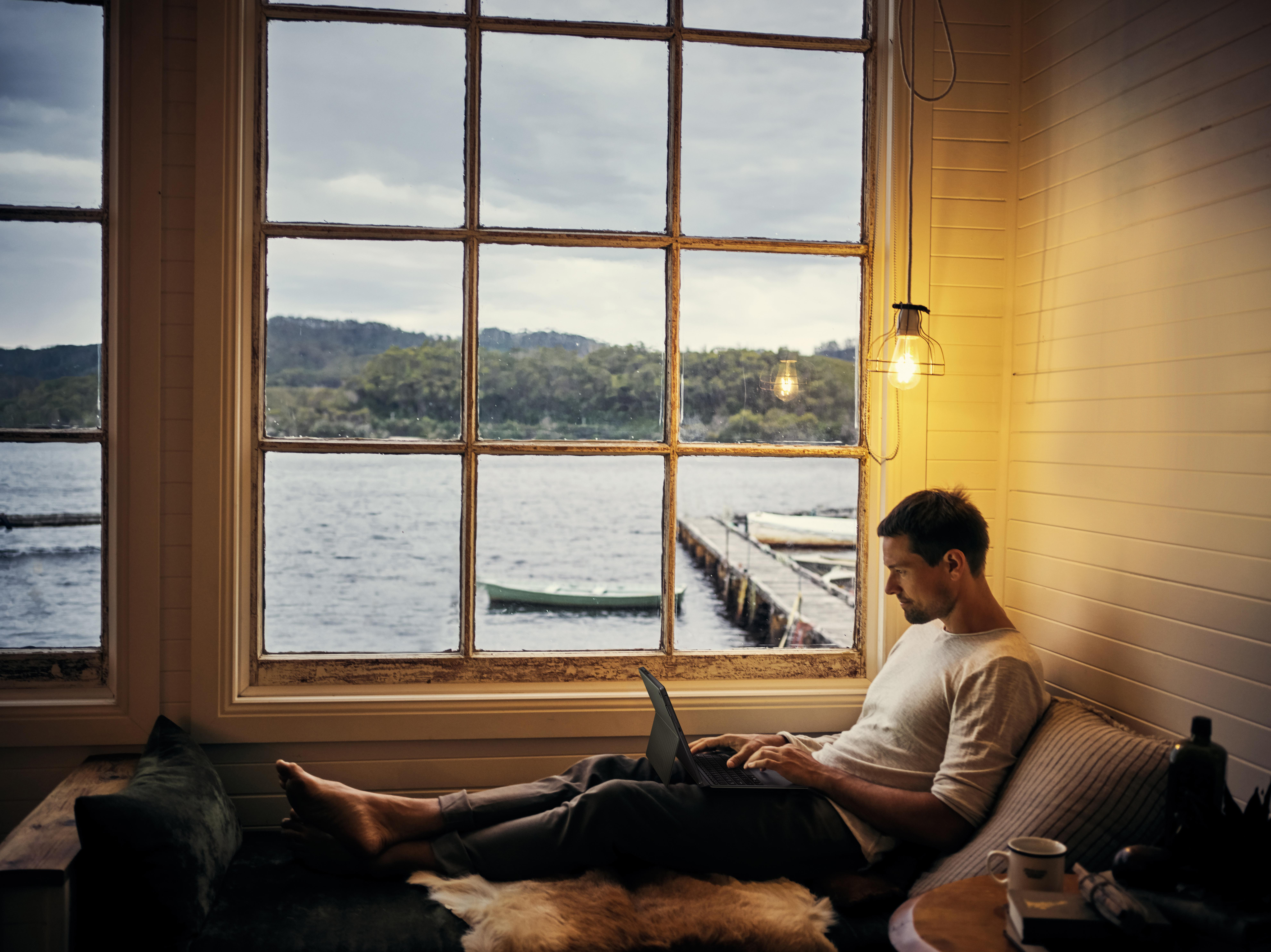 Mann mit Laptop am Fenster, blickt auf das Wasser hinaus.