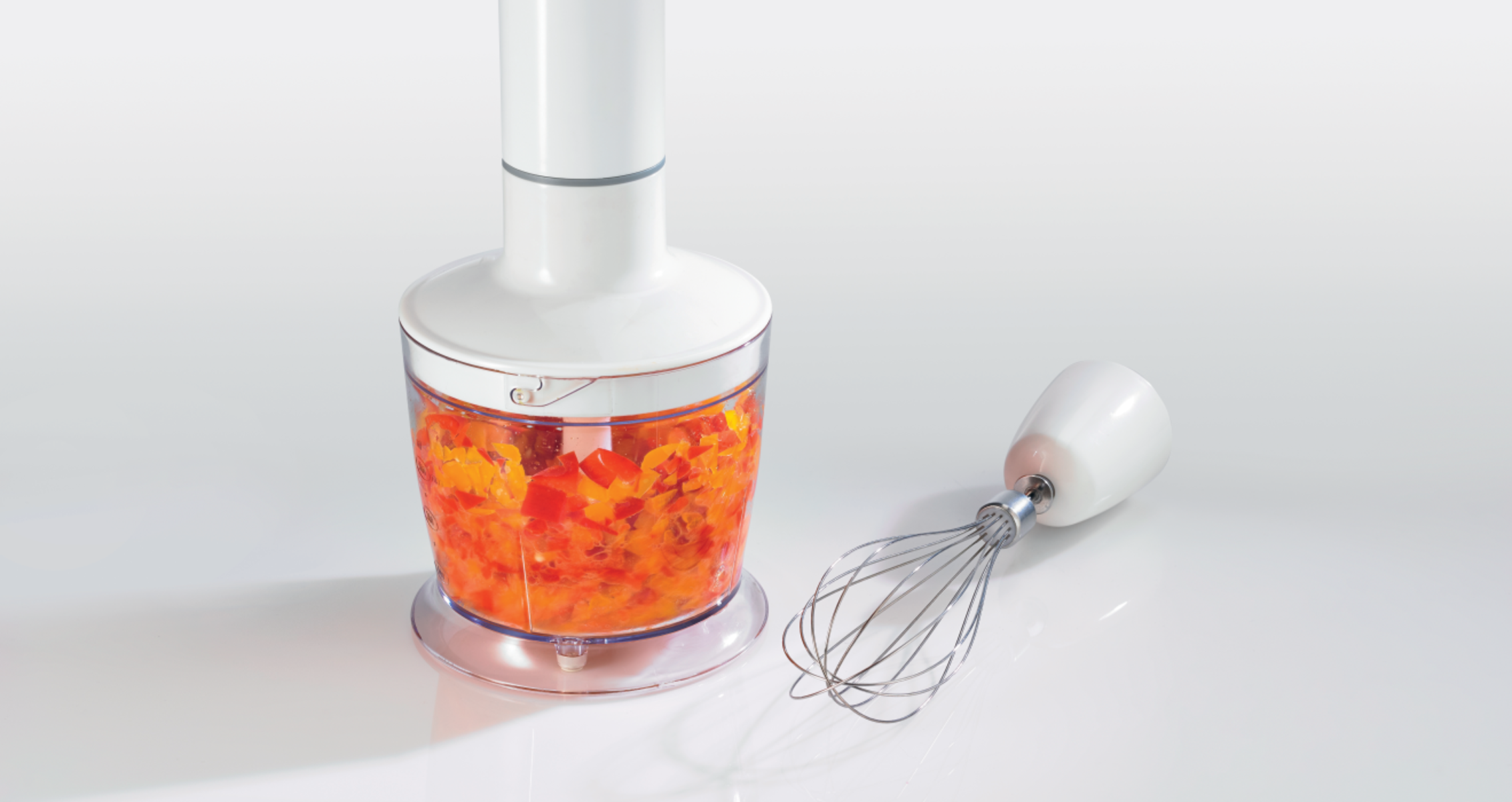 Kézi mixer egy átlátszó tartállyal, tele apróra vágott piros paprikával.