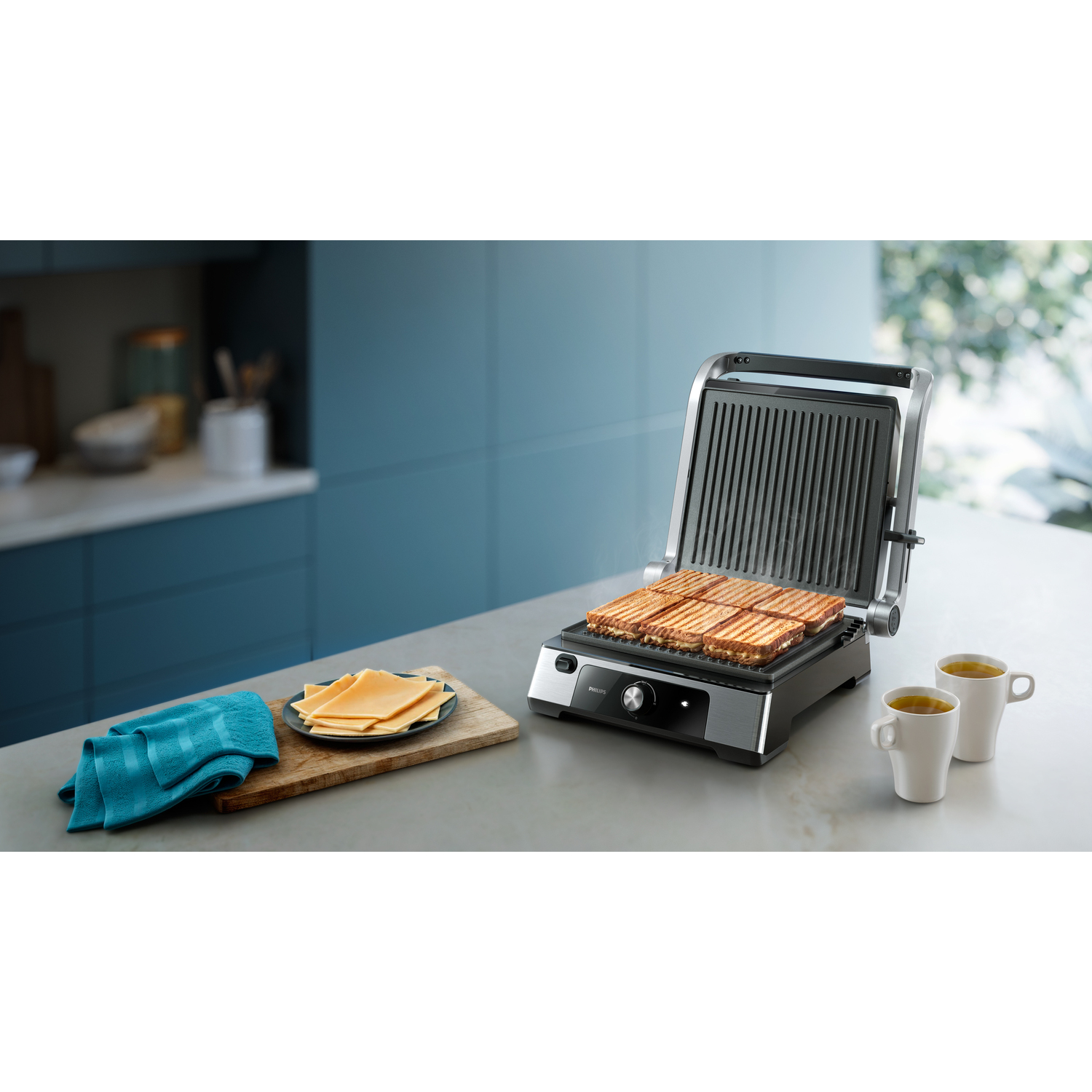 Sandwiches op een grill, houten snijplank met kaas en twee kopjes thee. Een blauwe handdoek ligt in de buurt.