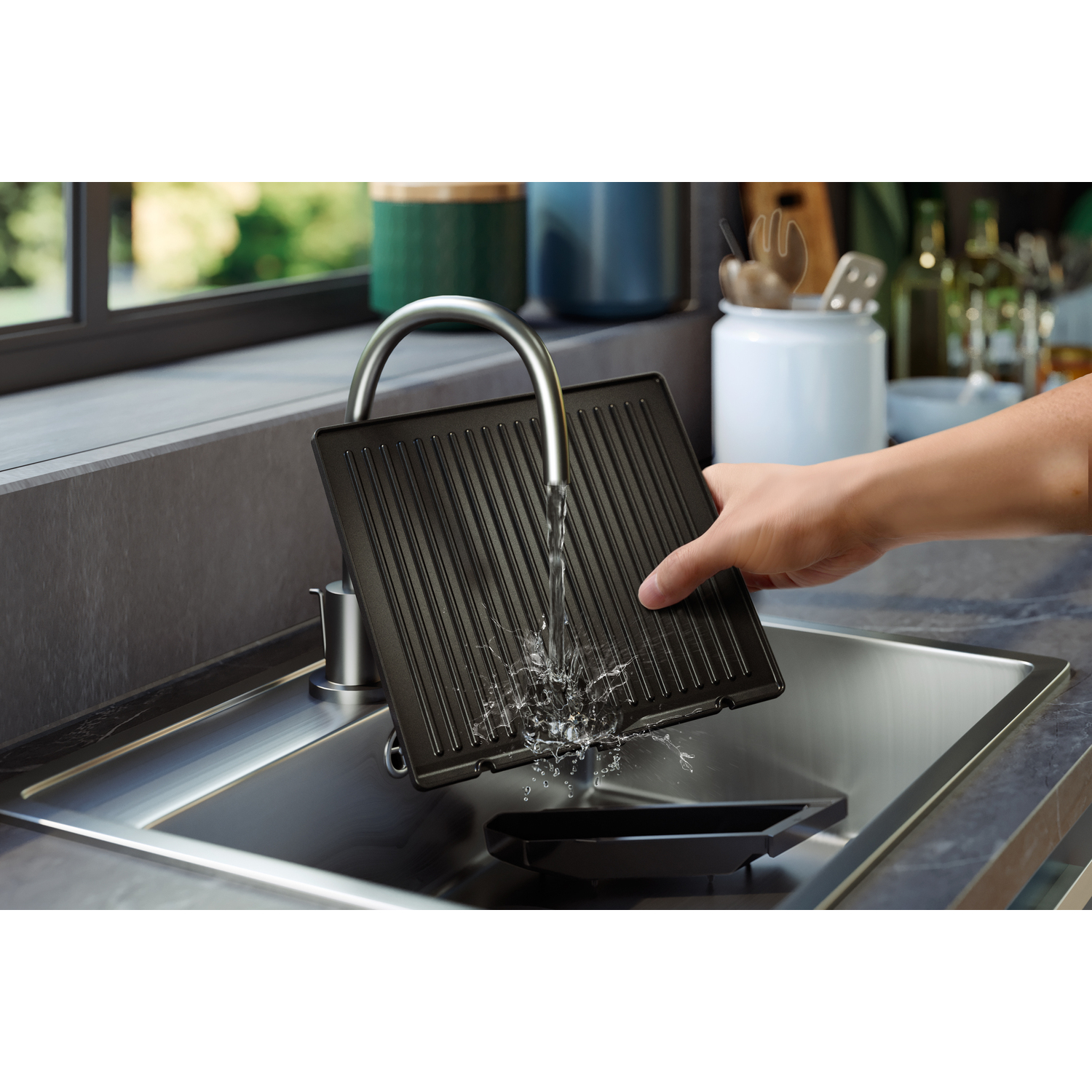 Une personne lave une plaque de cuisson noire dans un évier en acier inoxydable avec de l'eau courante.