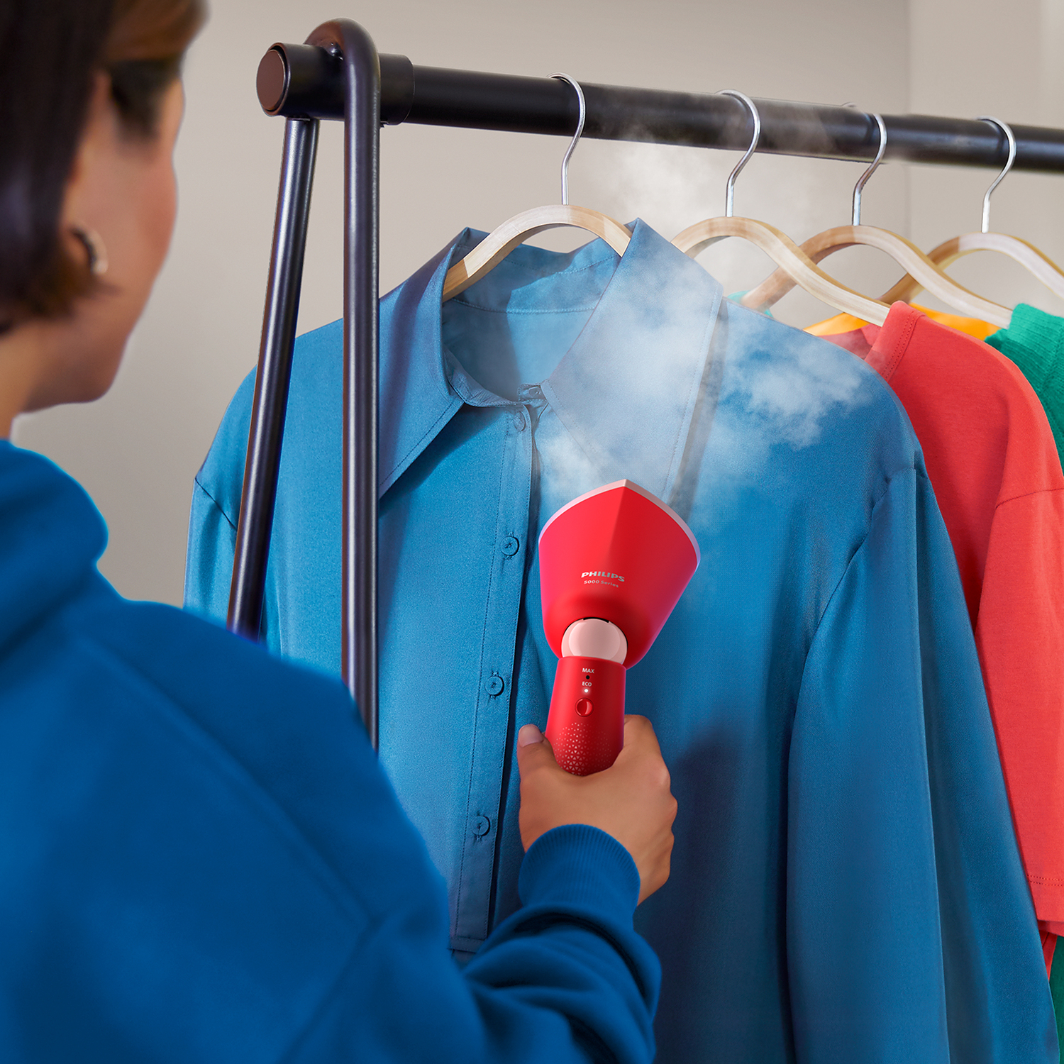 Woman steaming a blue shirt with a red Philips steamer, clothes on hangers.