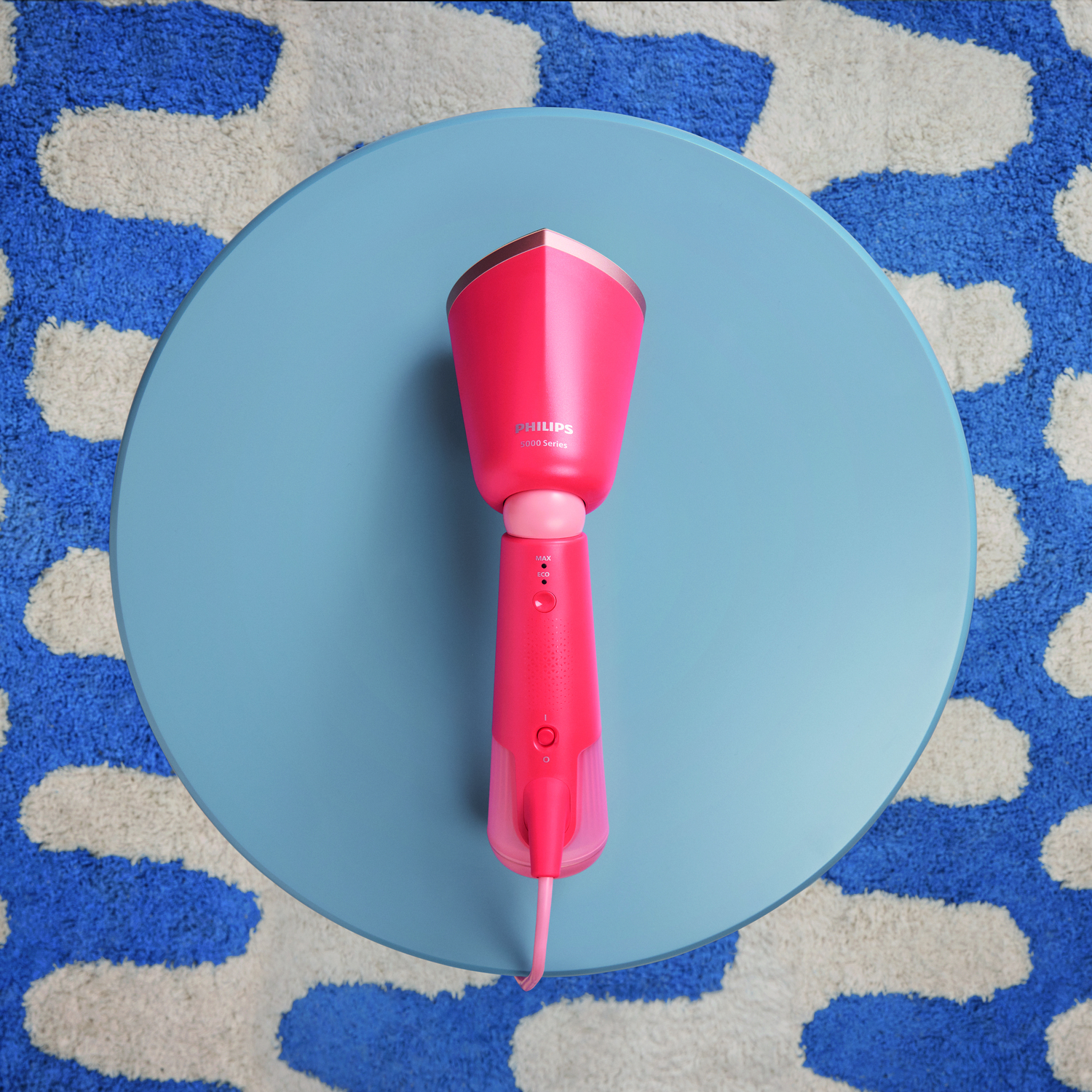 Pink and gold handheld garment steamer on a blue table. A patterned blue and white rug is below.