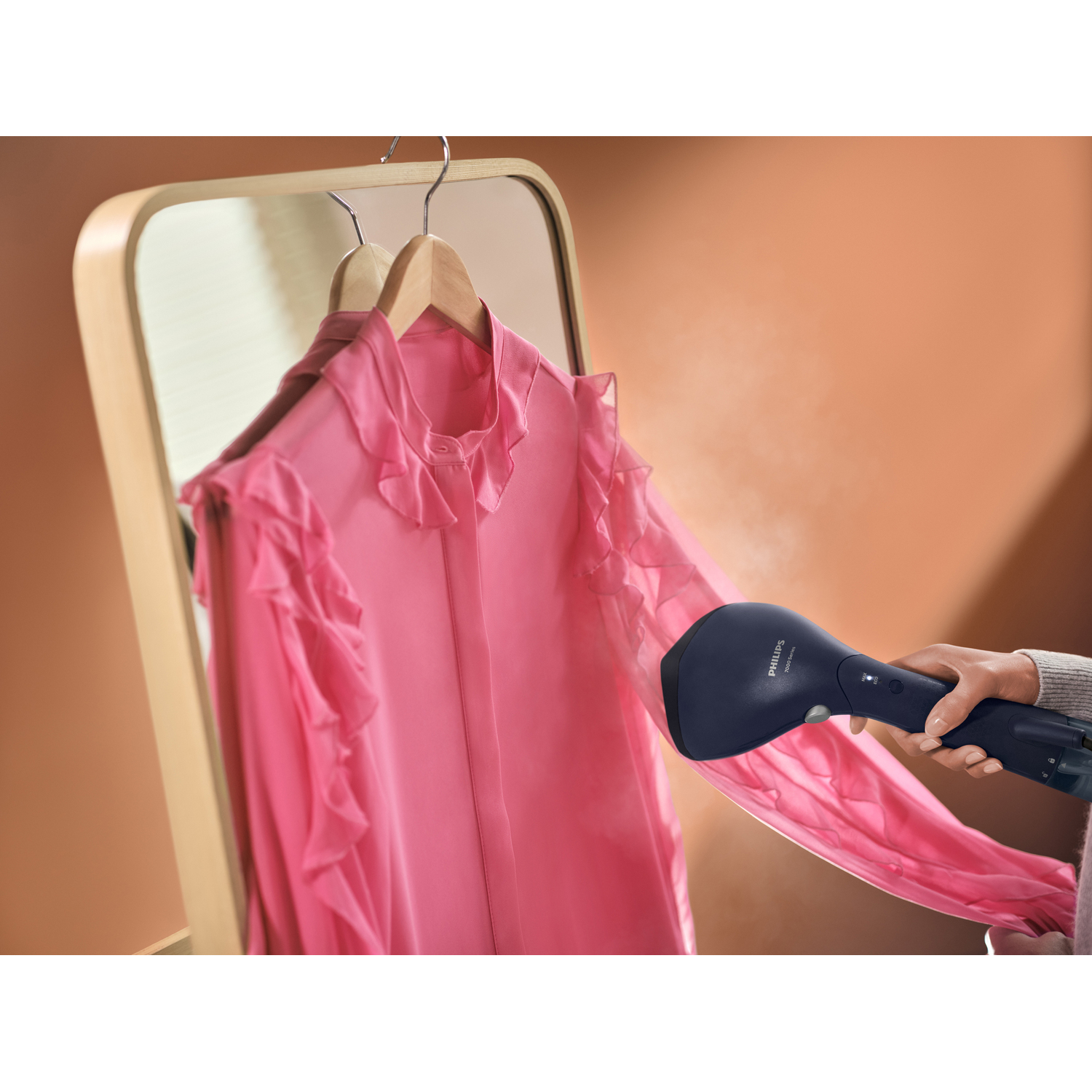 A pink shirt is being steamed in front of a mirror using a blue handheld steamer.