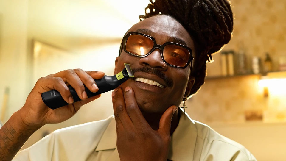 Mężczyzna przycinający brodę trymerem, uśmiechnięty, w okularach.