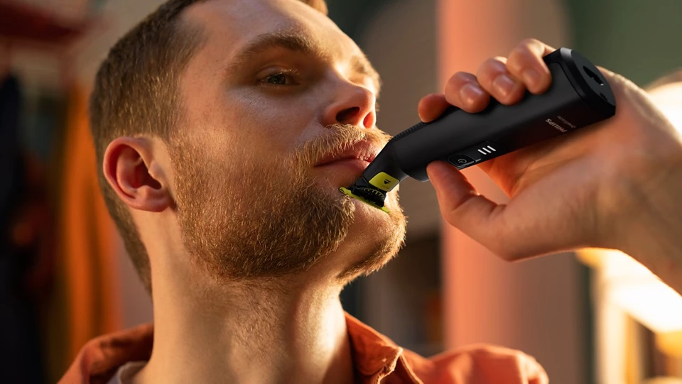 Mężczyzna goli brodę czarno-żółtą trymerem trzymanym przez inną osobę.