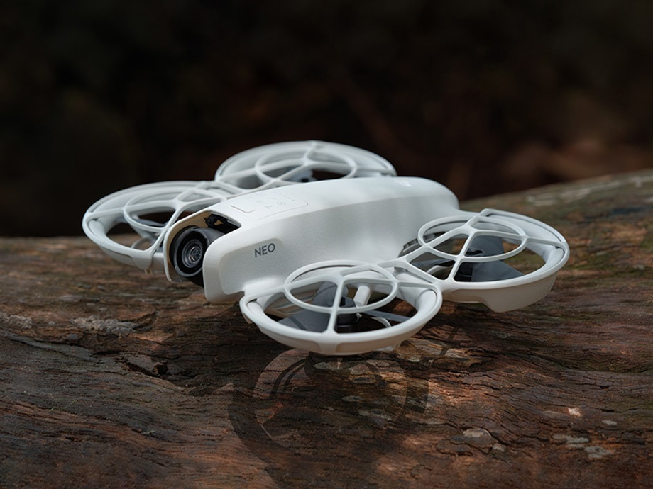 Biały dron na kłodzie, z czterema śmigłami. Dron jest oznaczony jako NEO.