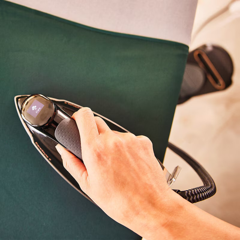 Osoba prasuje ciemnozielony materiał żelazkiem. Żelazko jest czarne.