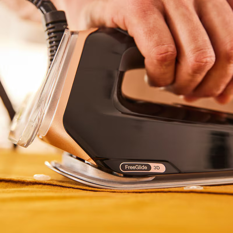 Osoba prasująca żółtą tkaninę. Używana jest czarno-złota żelazko, z ustawieniem na dole.