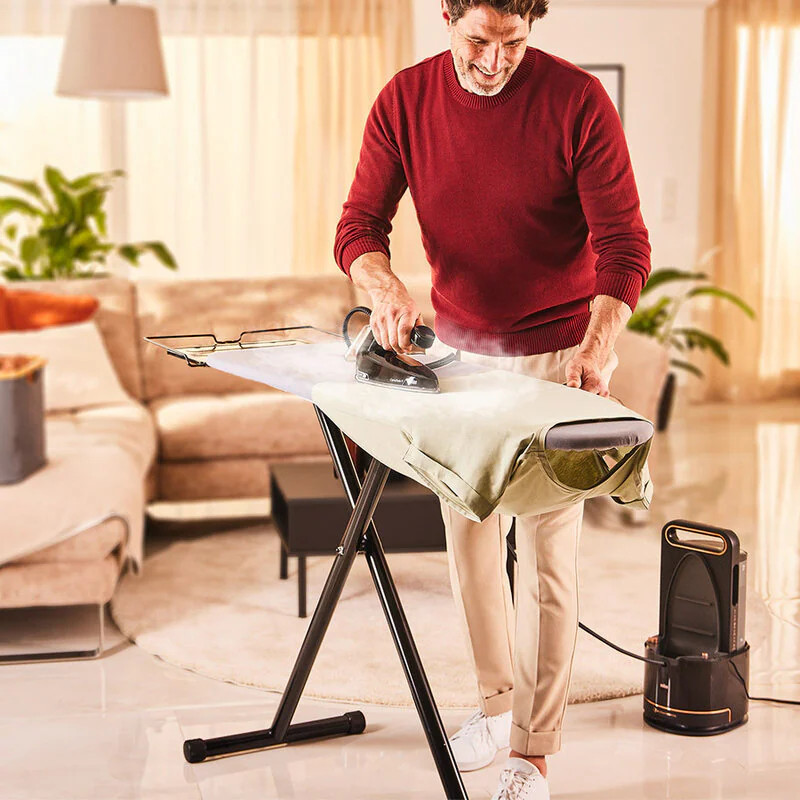 Mężczyzna prasuje ubrania na desce do prasowania w salonie. Ma na sobie czerwony sweter i białe spodnie.