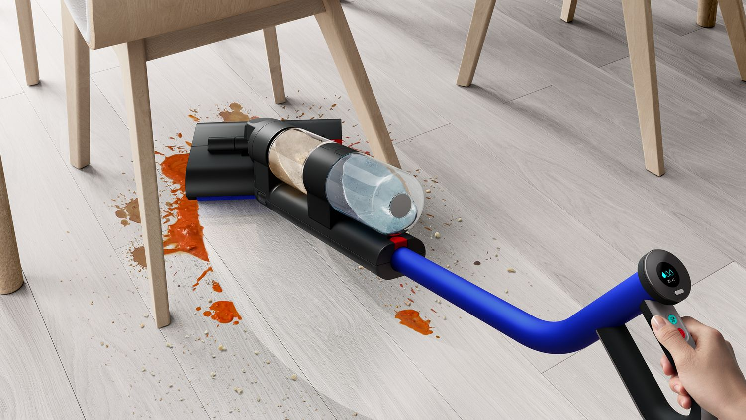 Un aspirateur nettoie un sol taché. Il y a des chaises et une table à proximité.