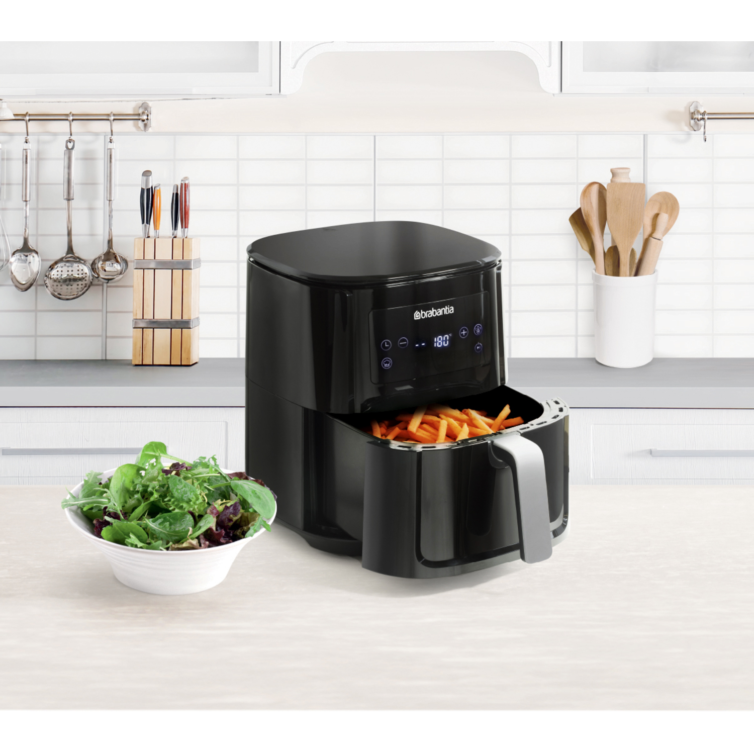 Black air fryer with a bowl of salad and fries inside. Kitchen setting with white tile background.
