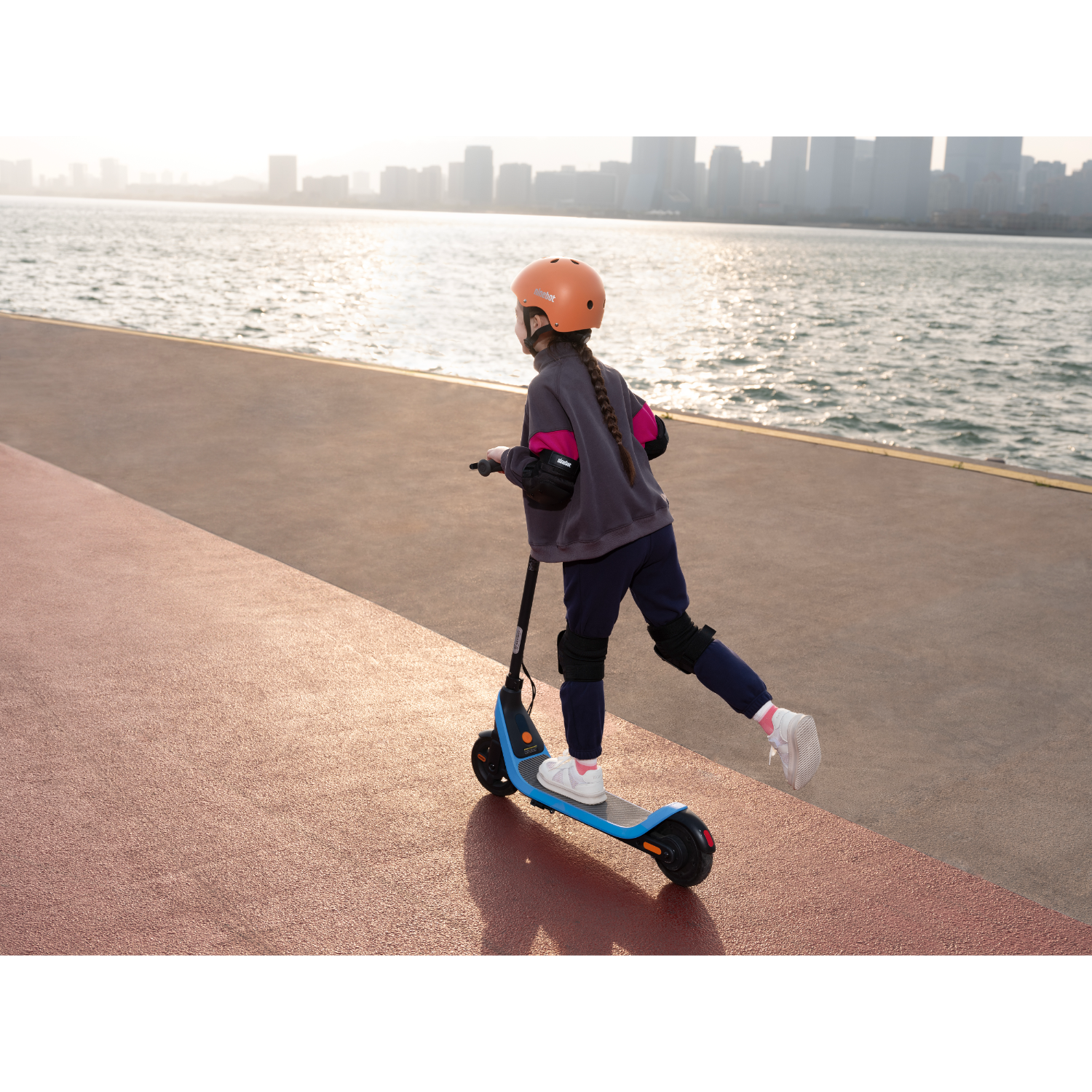 Girl on scooter, orange helmet, on a red path near water, city skyline background.