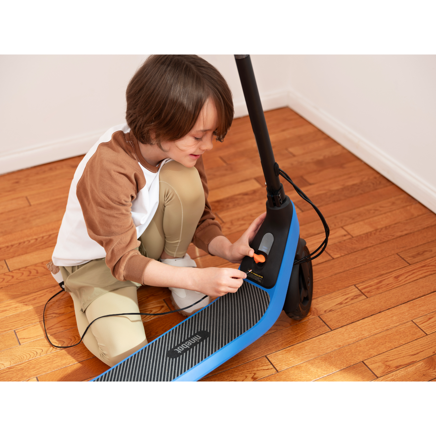Child charging a blue and black electric scooter. Wooden floor, white wall.