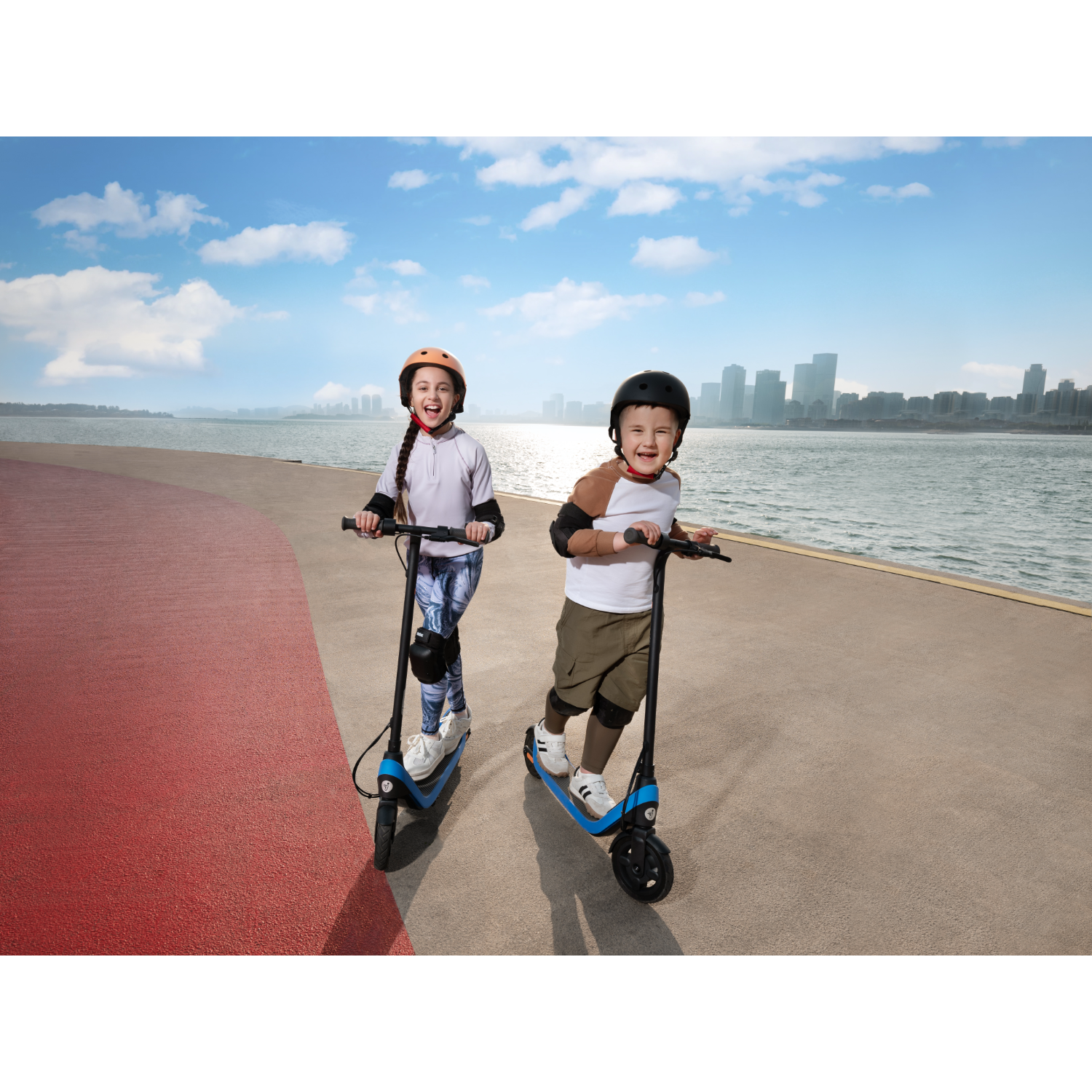 Two kids on scooters smiling. Blue sky, city in background, and red path.