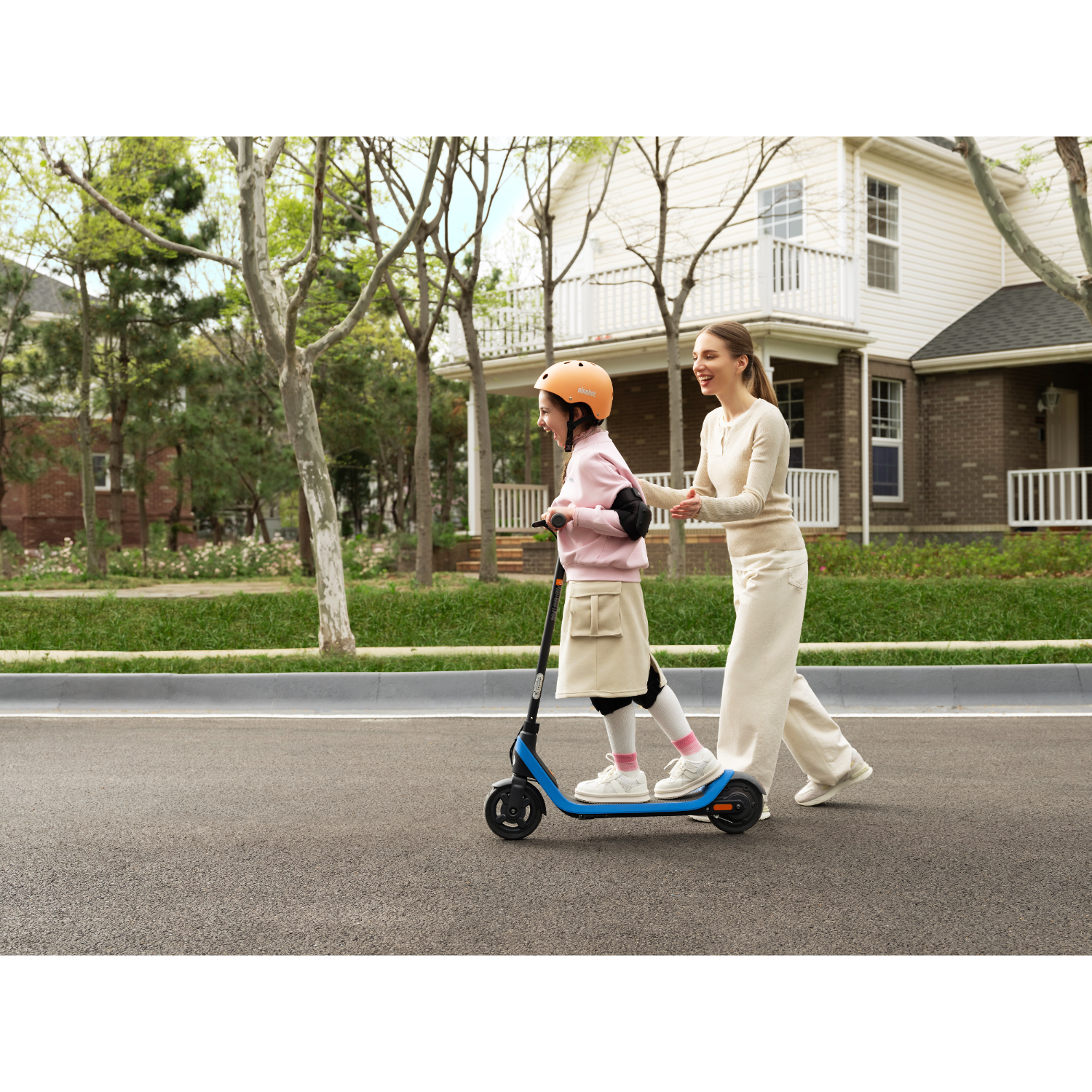 A mother assists a child riding a scooter on a road in front of a house.