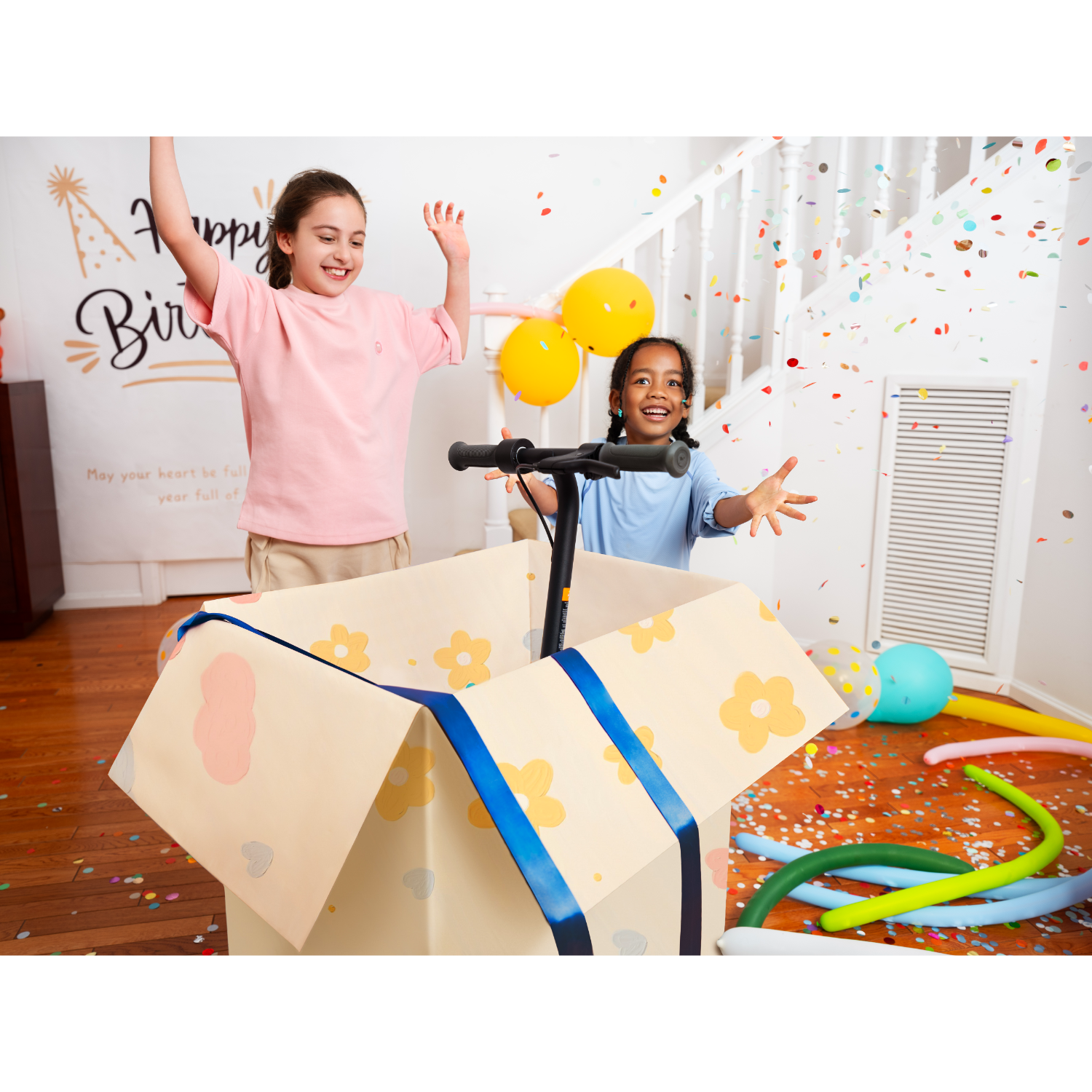 Two young girls in a room with balloons and confetti, one in a box with a scooter.