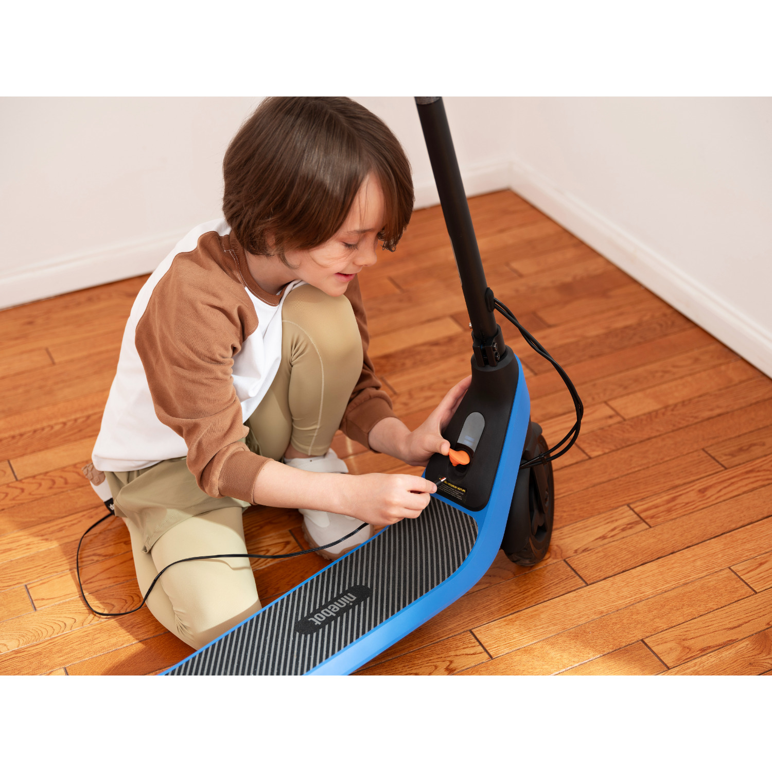 Een jongen knielt, sluit een kabel aan op een blauw-zwarte scooter, houten vloer.