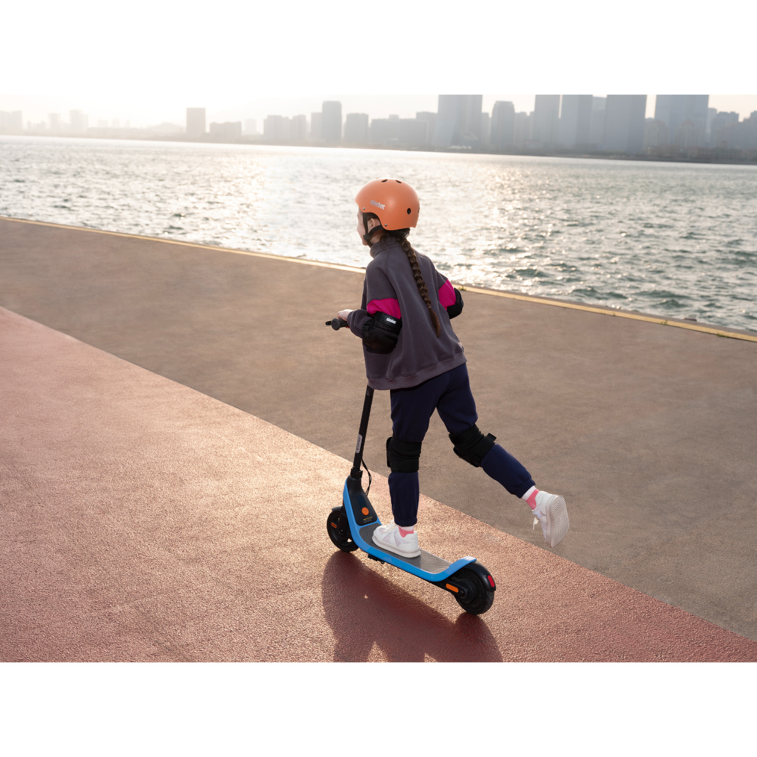Een meisje op een blauw-zwarte scooter op een rood oppervlak, met een skyline op de achtergrond.