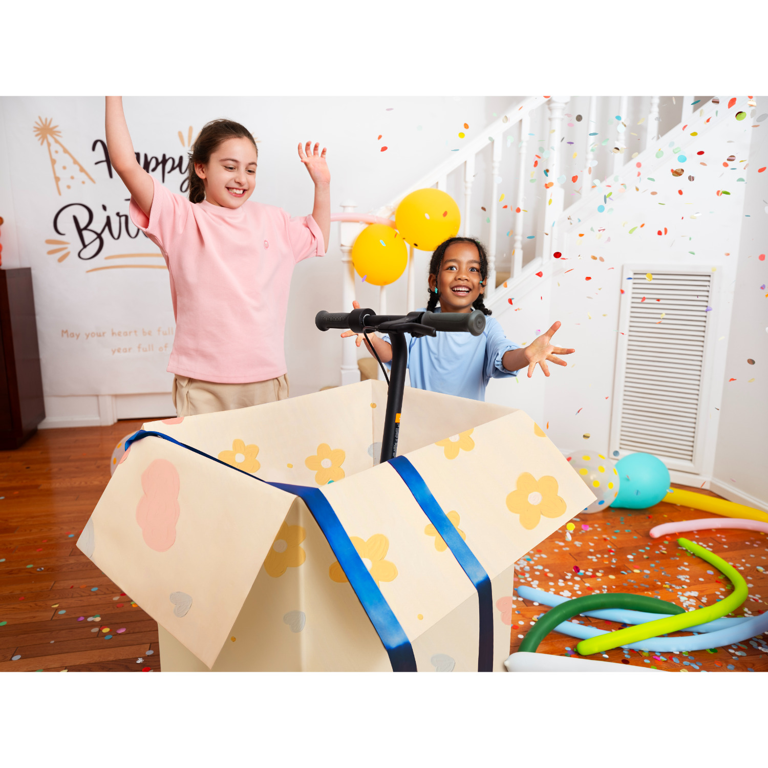 Twee meisjes vieren. Een scooter in een kartonnen doos vol ballonnen en confetti.