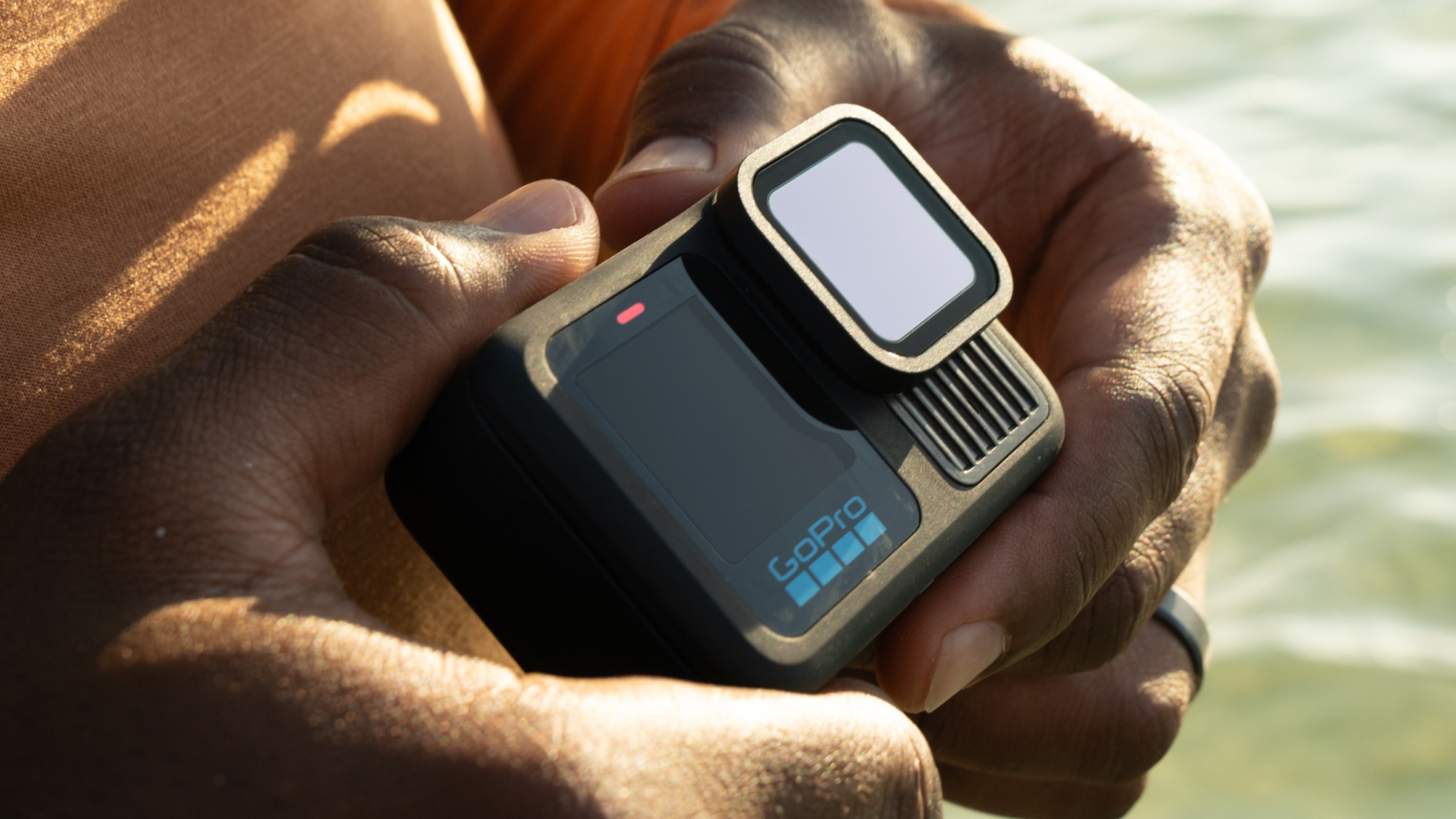 Eine Person hält eine schwarze GoPro-Kamera mit hochklappbarem Bildschirm im Freien am Wasser.