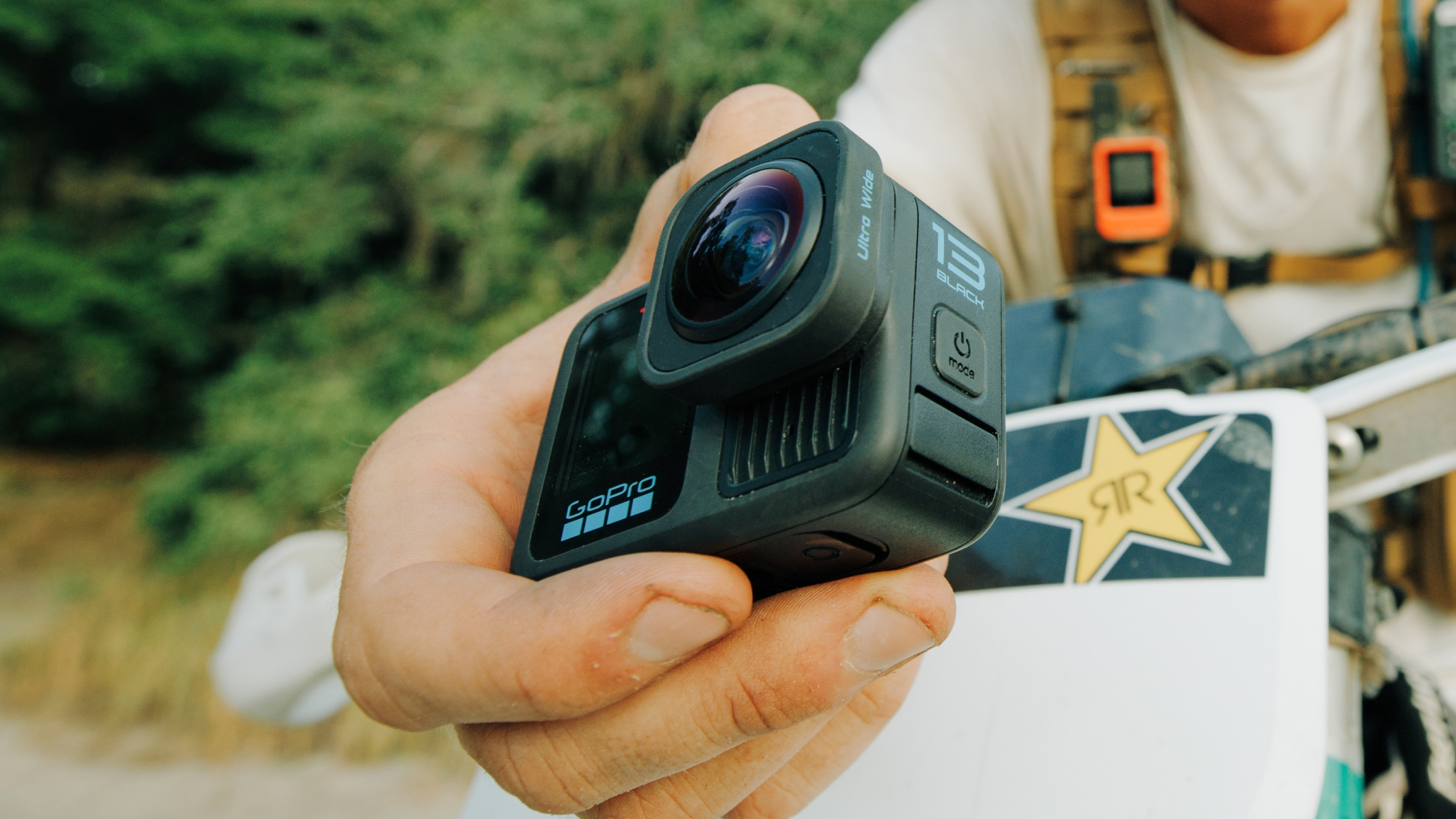 Eine GoPro-Kamera wird von einer Person vor grünen Bäumen und einer weißen Oberfläche gehalten.