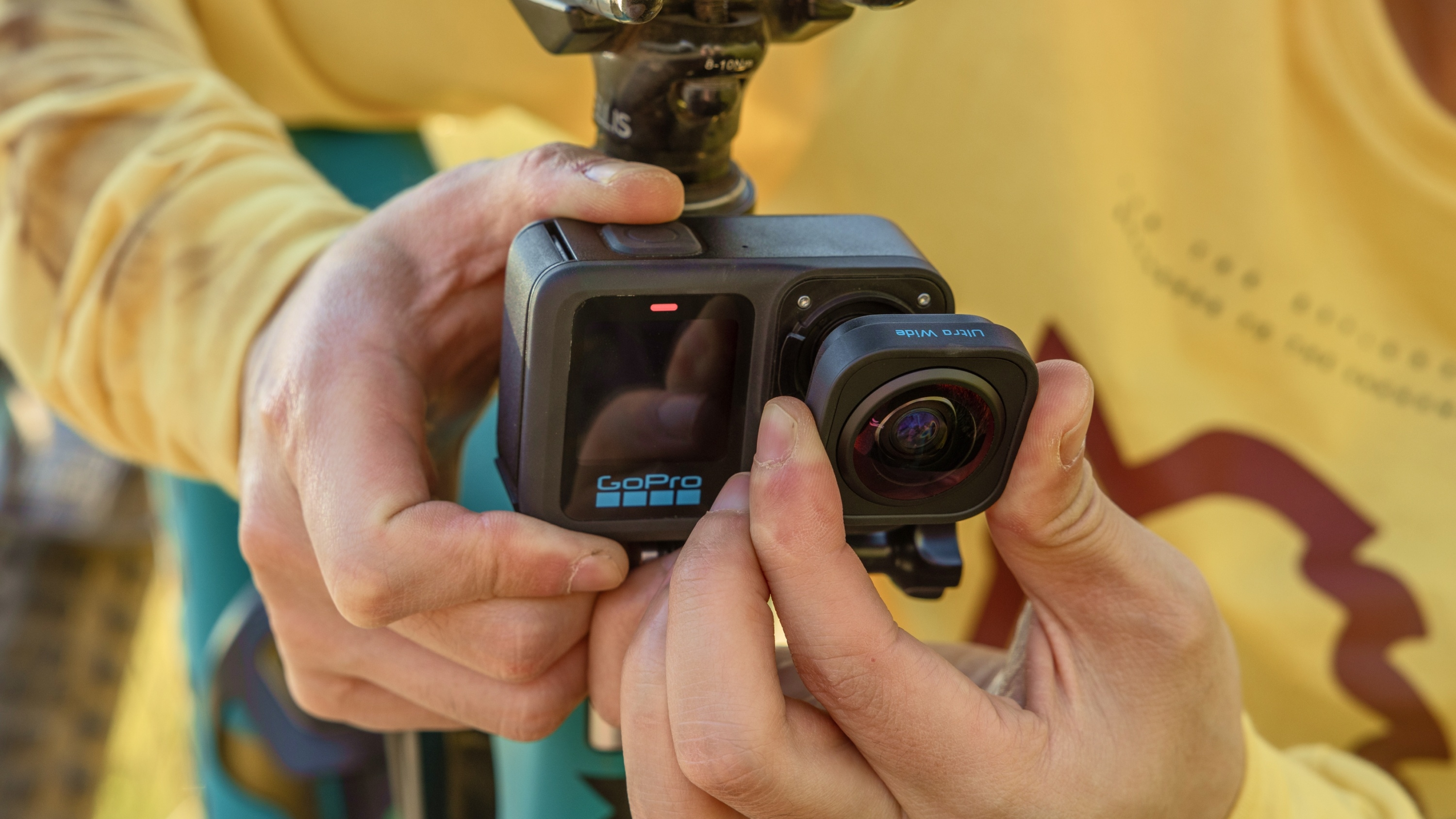 Person befestigt GoPro-Kameralinse an einer Halterung. Gelbes Hemd sichtbar.