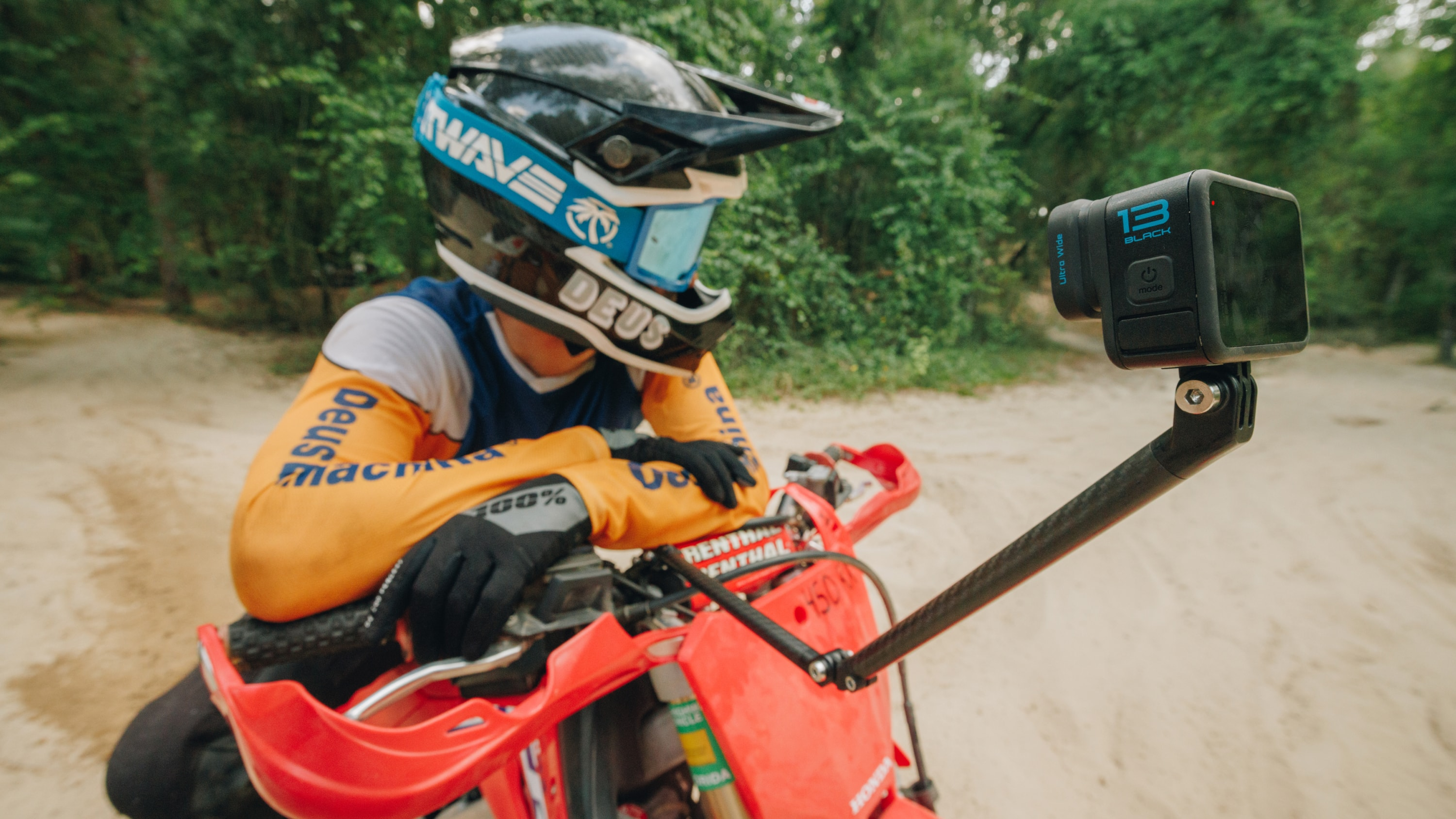 Motorradfahrer mit Helm und GoPro-Kamera an einem Selfie-Stick. Rotes Motorrad auf einem Feldweg im Wald.