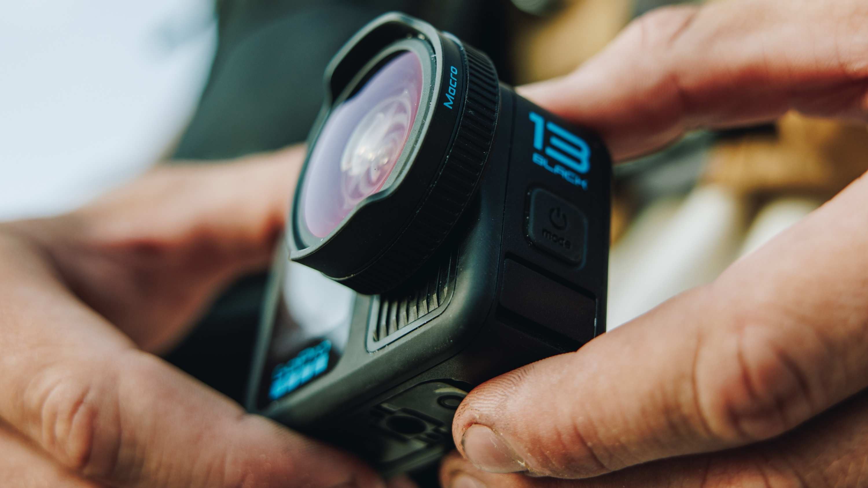 Schwarze GoPro-Kamera, gehalten von Händen, mit Makroobjektiv. Power- und Modus-Tasten sind sichtbar.