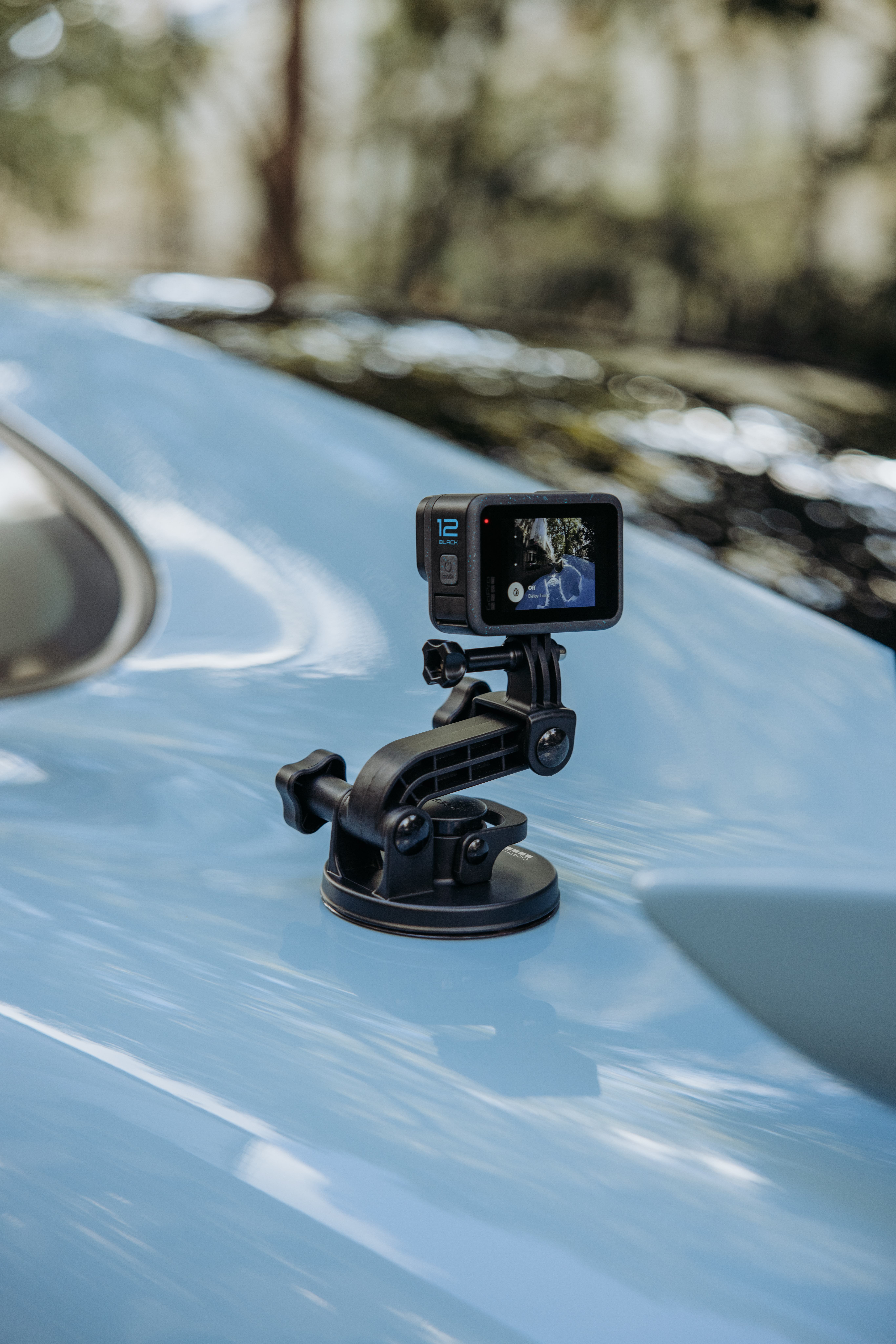 Une caméra GoPro noire est montée sur un toit de voiture bleu. L'écran affiche une scène. Le fond est des arbres flous.