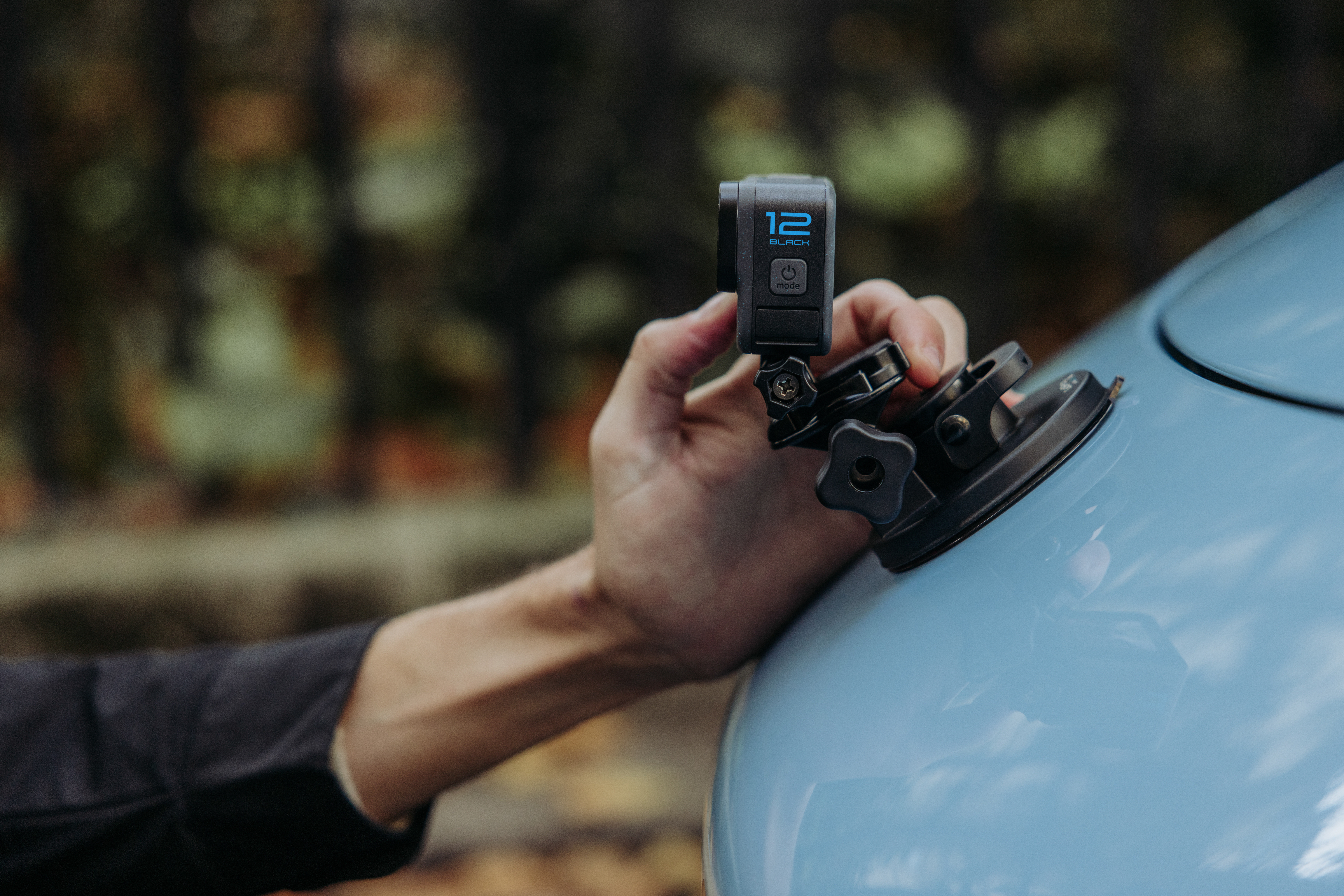 Une main tient une caméra GoPro fixée à une voiture. La caméra est noire avec du texte bleu. La voiture est bleu clair.