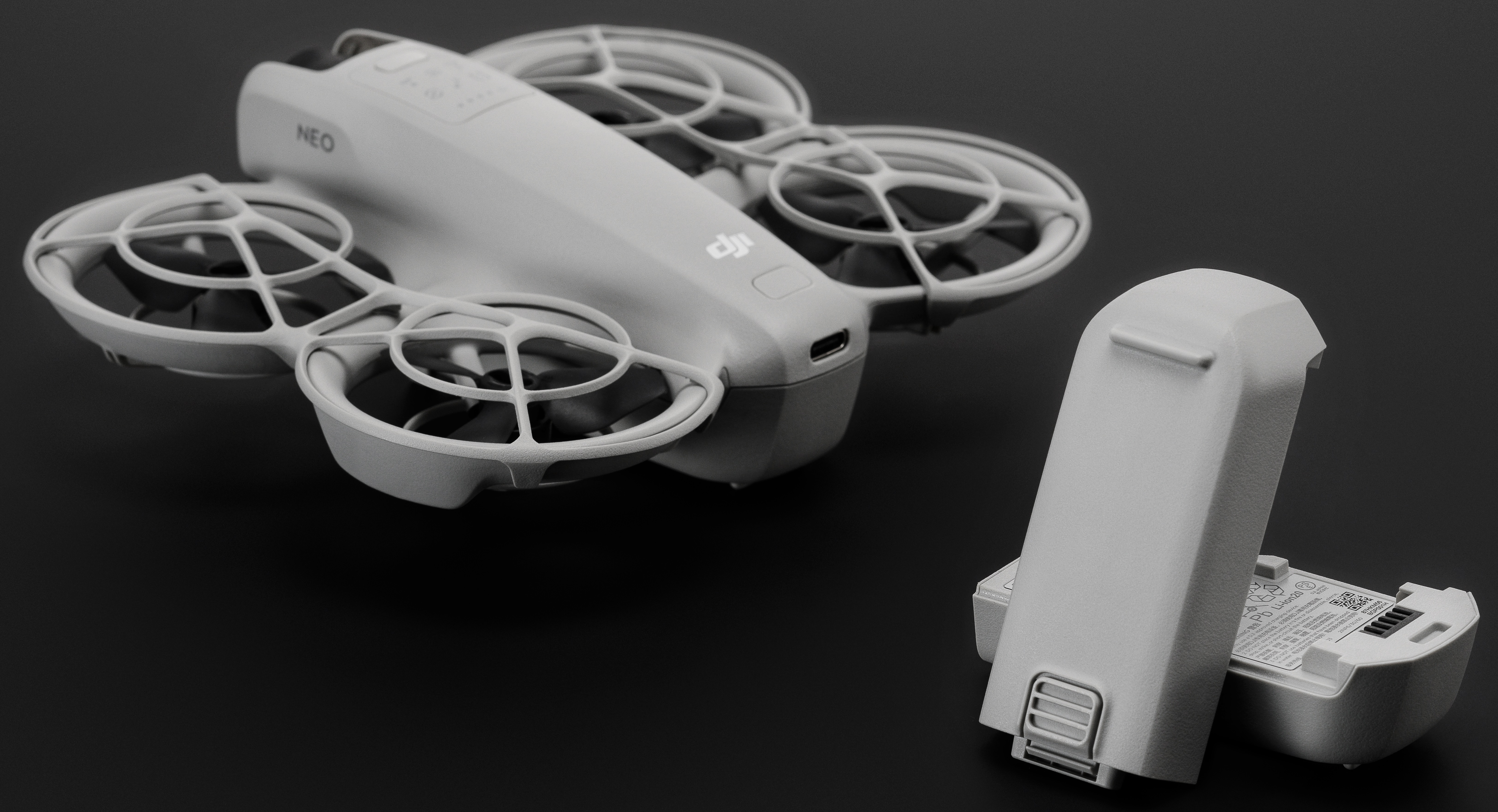 Biały dron z czterema śmigłami i baterią obok, na czarnym tle.