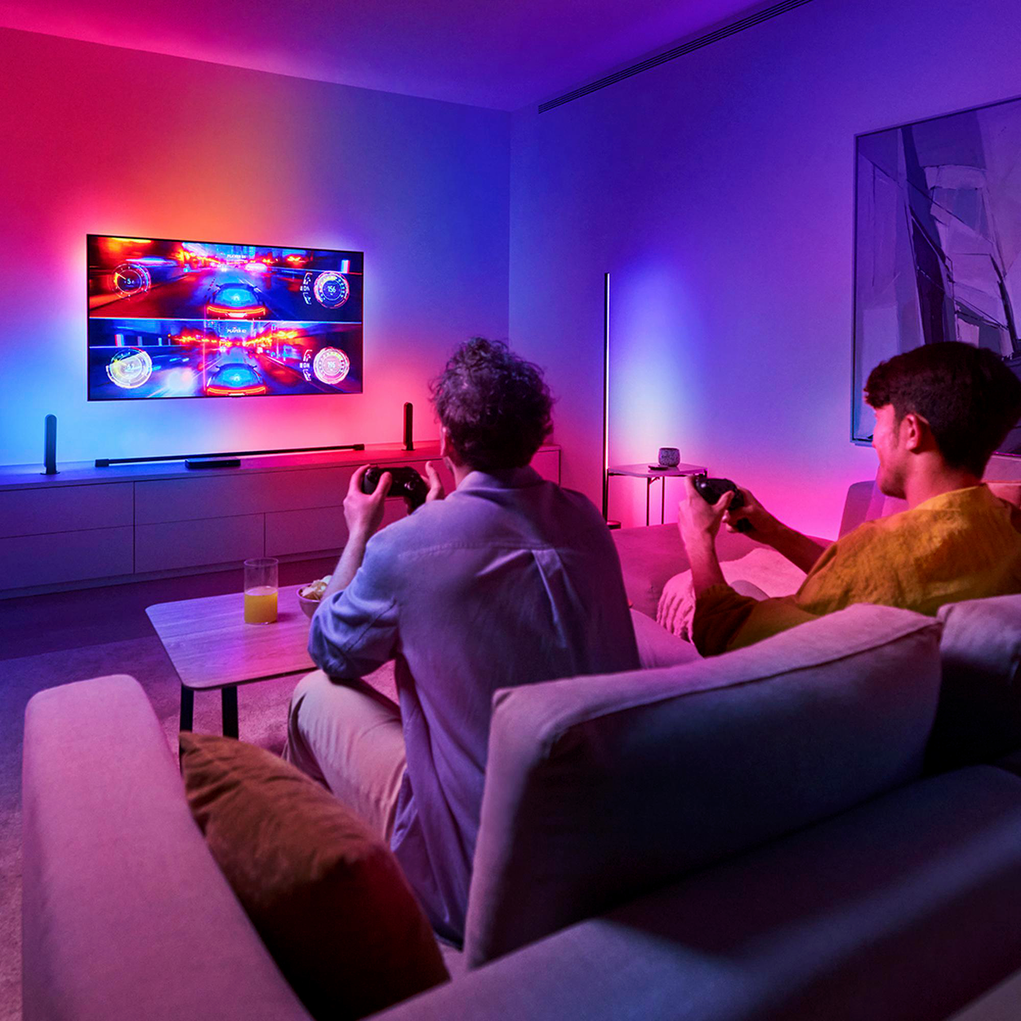 Zwei Männer spielen Videospiele auf einem Sofa. Fernseher zeigt Rennspiel. Farbige Lichter beleuchten den Raum.