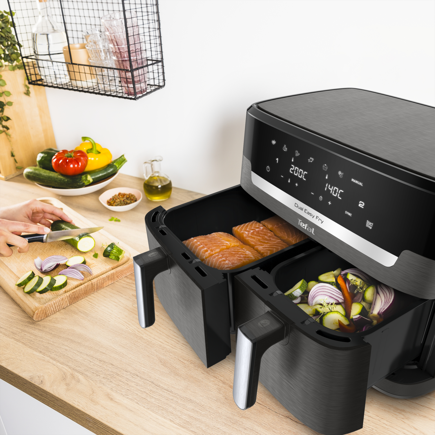 An air fryer shows salmon and vegetables in its baskets. A person prepares vegetables on a cutting board.