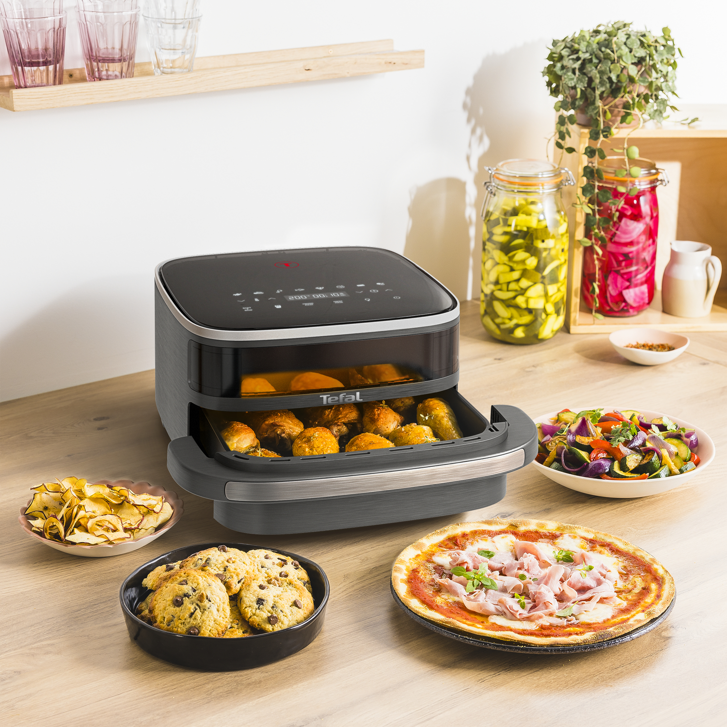 An air fryer with cooked food, cookies, pizza, and snacks. Jars and plants are in the background.