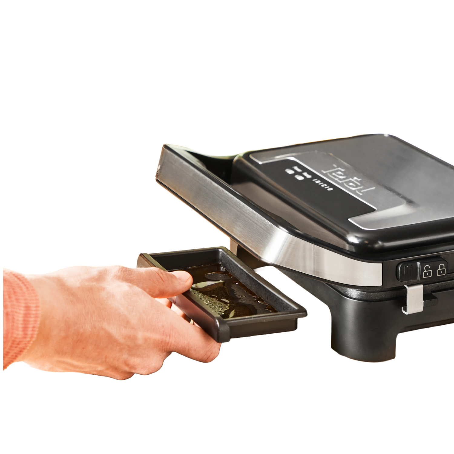 Een hand houdt een zwarte bak met olie vast, naast een zwart-zilveren Tefal grill. De achtergrond is wazig en bruin.