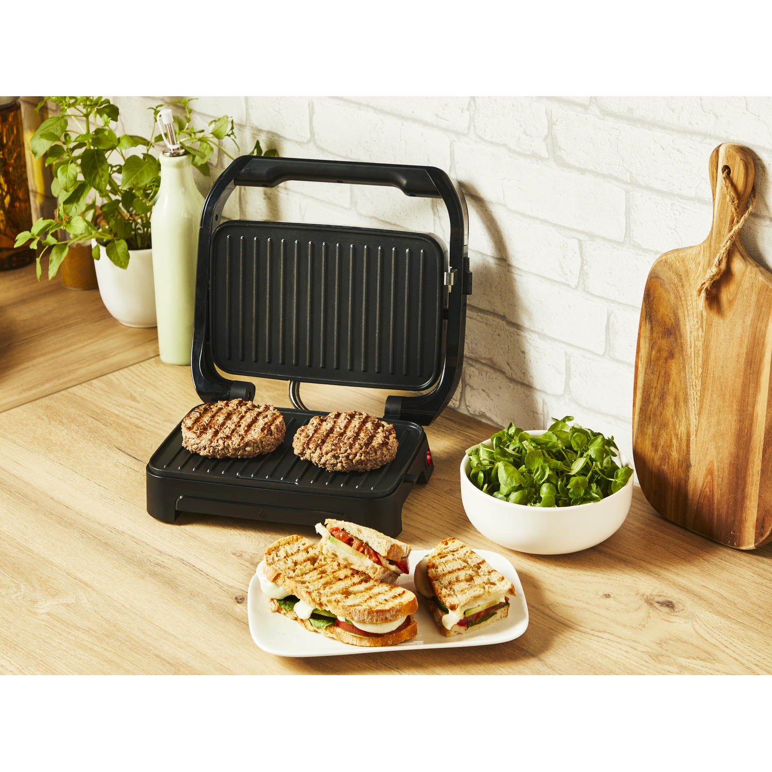 Sandwiches en burgers op een grill, met een grillpers. Groene salade en houten plank.