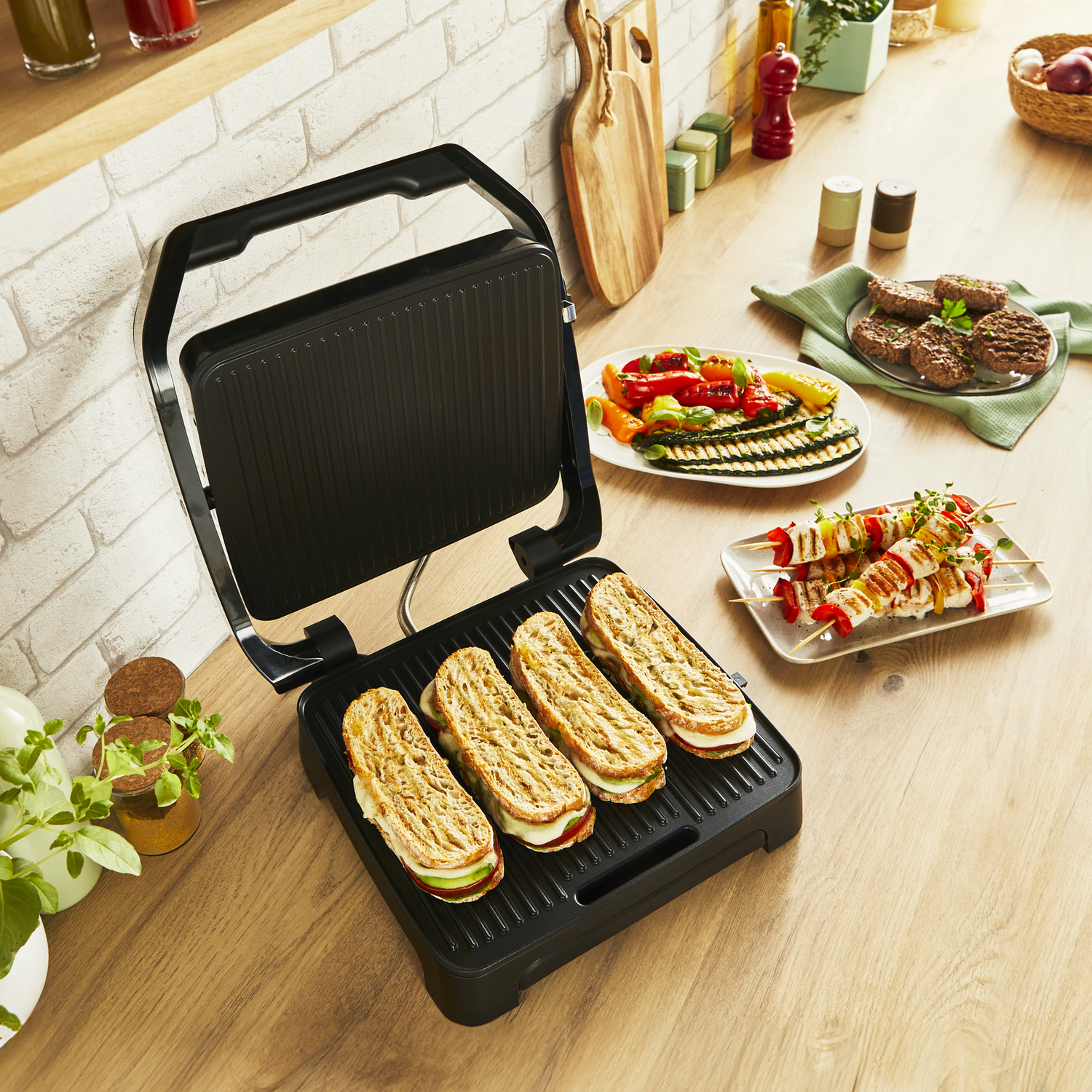 A griddle with sandwiches is open. Plates of grilled vegetables and skewers are on the counter.