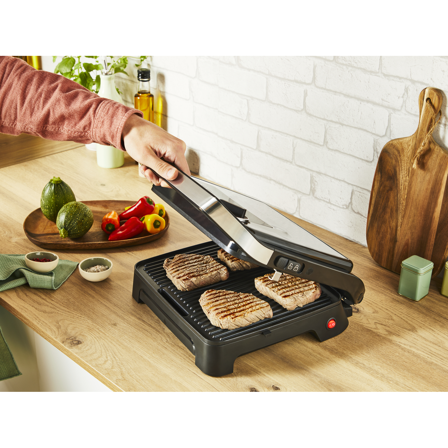 A person grilling steaks with a black grill on a wooden surface, with vegetables nearby.