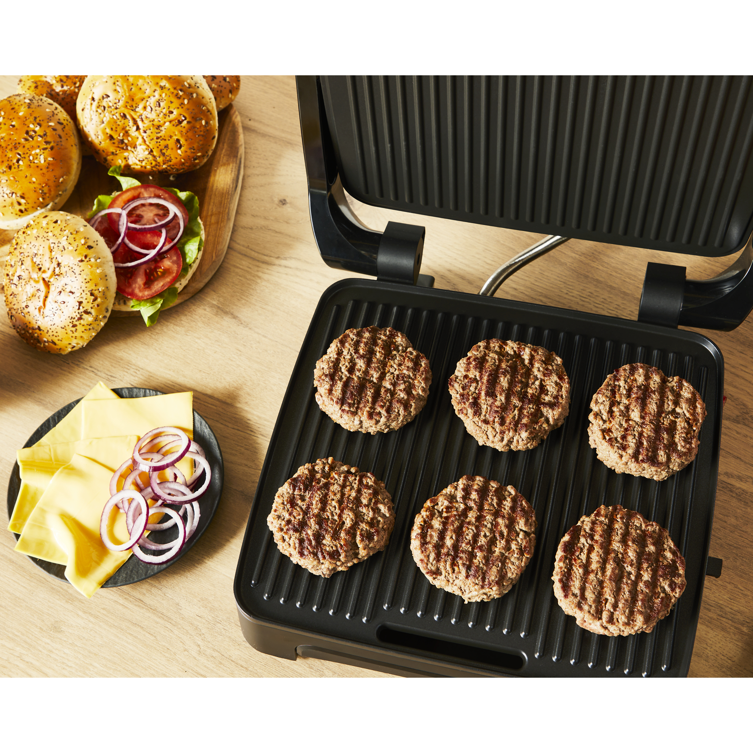 A black grill with cooked burgers, cheese, sliced onions, and buns on a wooden surface.