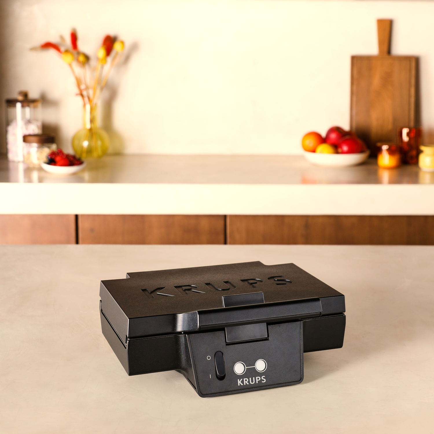 A black waffle maker is on a counter, with fruit and ingredients in the background.