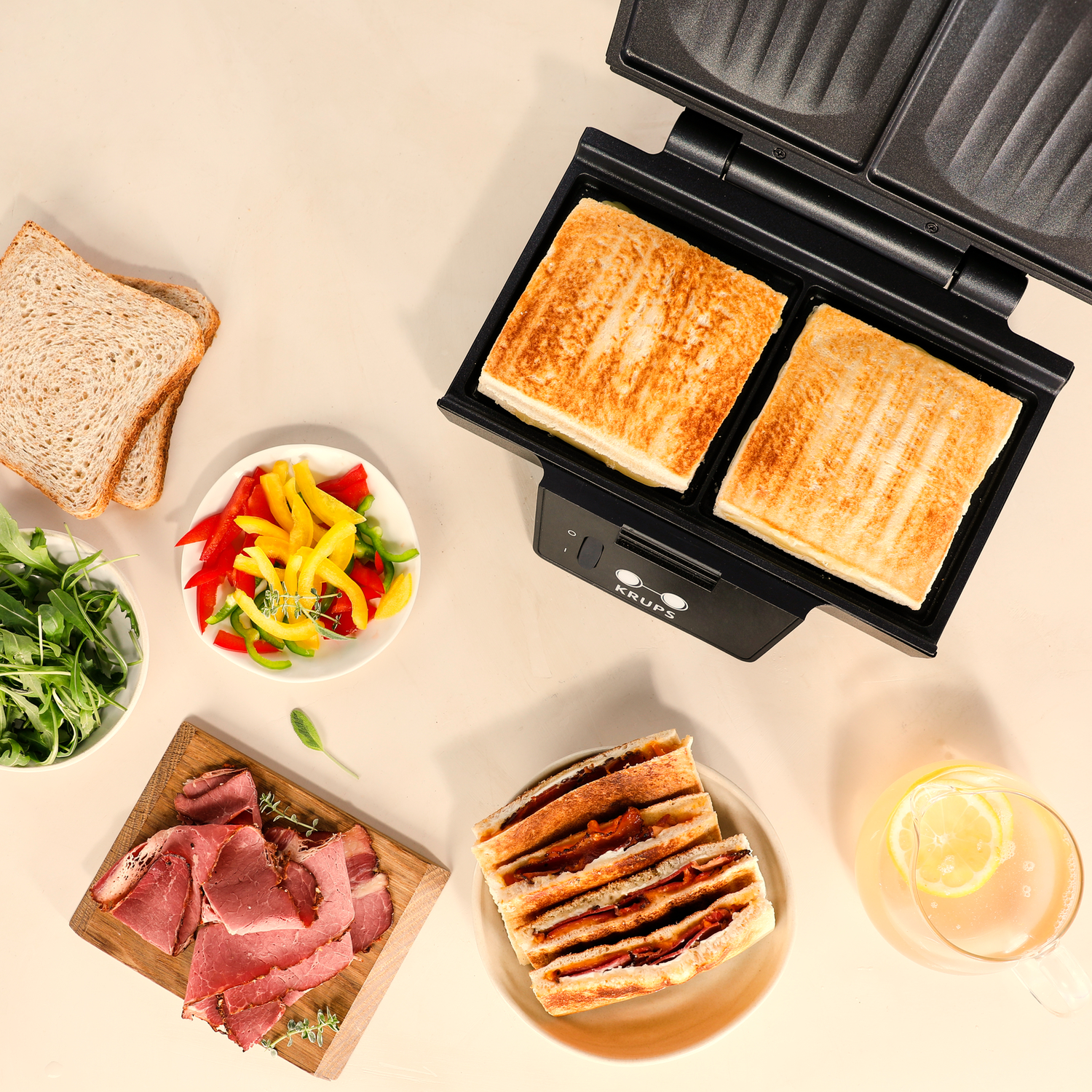 Sandwiches in a sandwich maker with side dishes on a light surface.