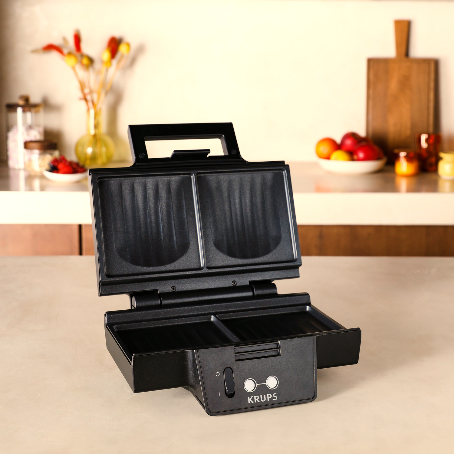 An open black Krups waffle maker on a light surface, with a blurred kitchen background.
