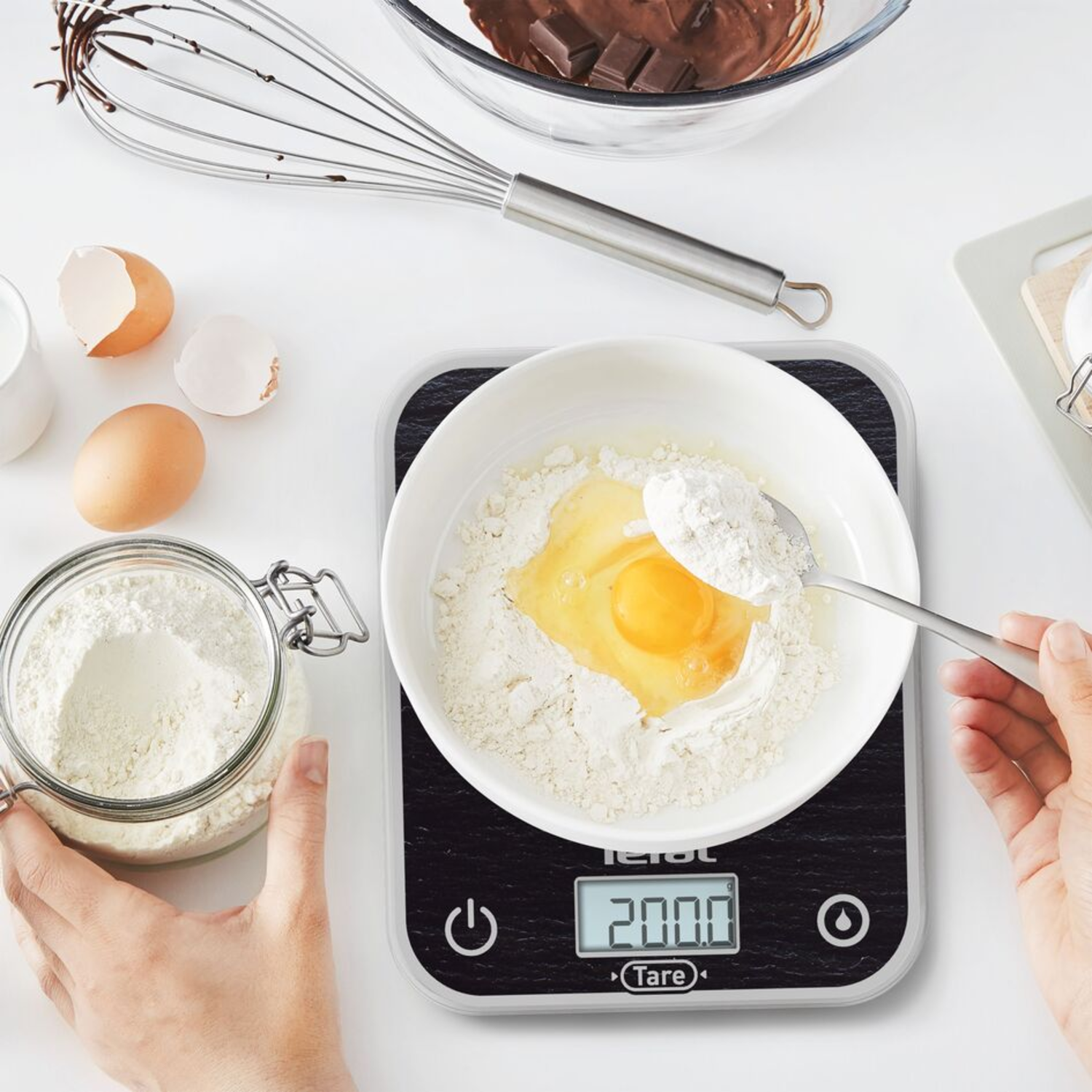 Ingredients for baking: flour in a bowl with an egg, spoon with flour, jar with flour, eggs, whisk, chocolate.