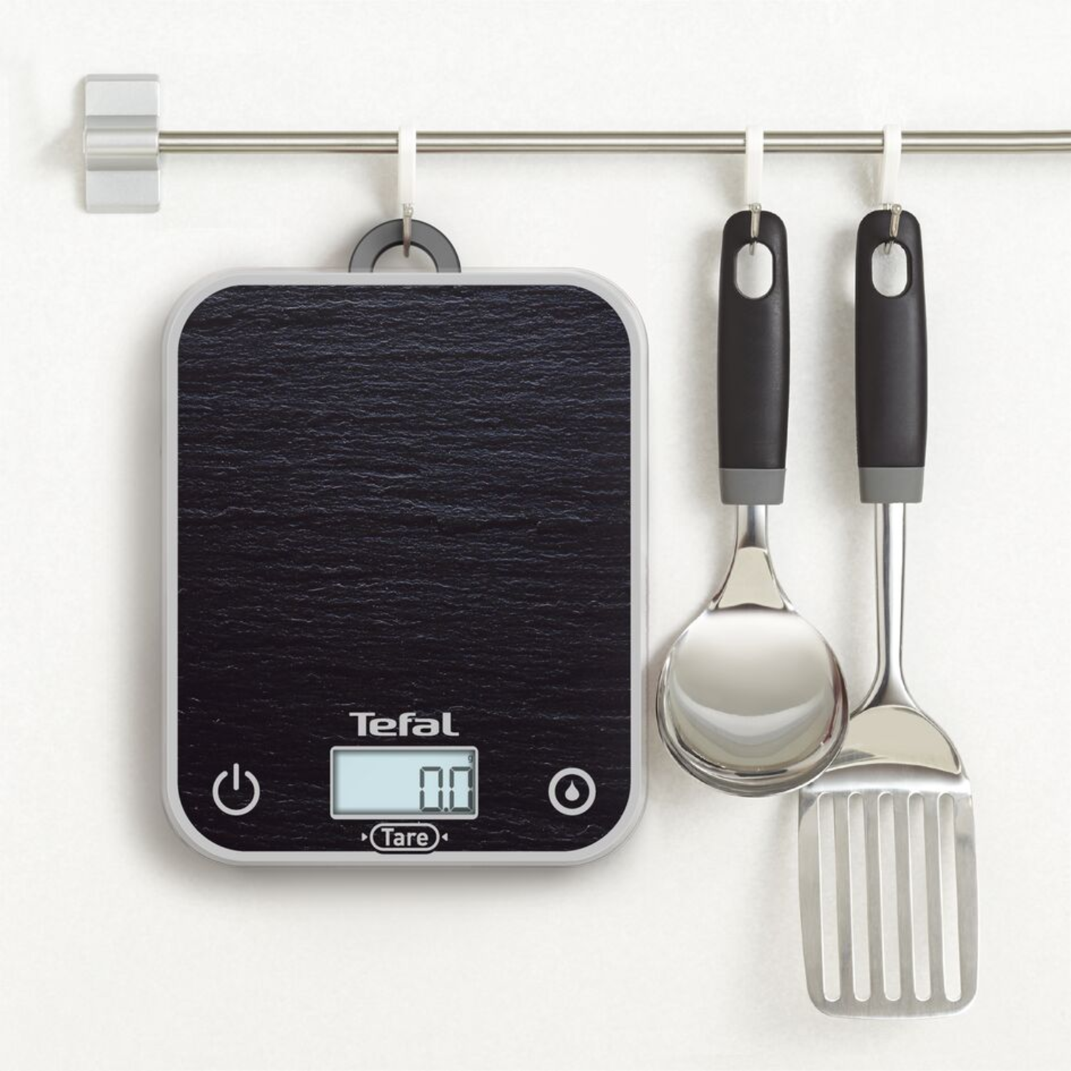 Tefal kitchen scale with two utensils hanging on a metal bar, white wall.